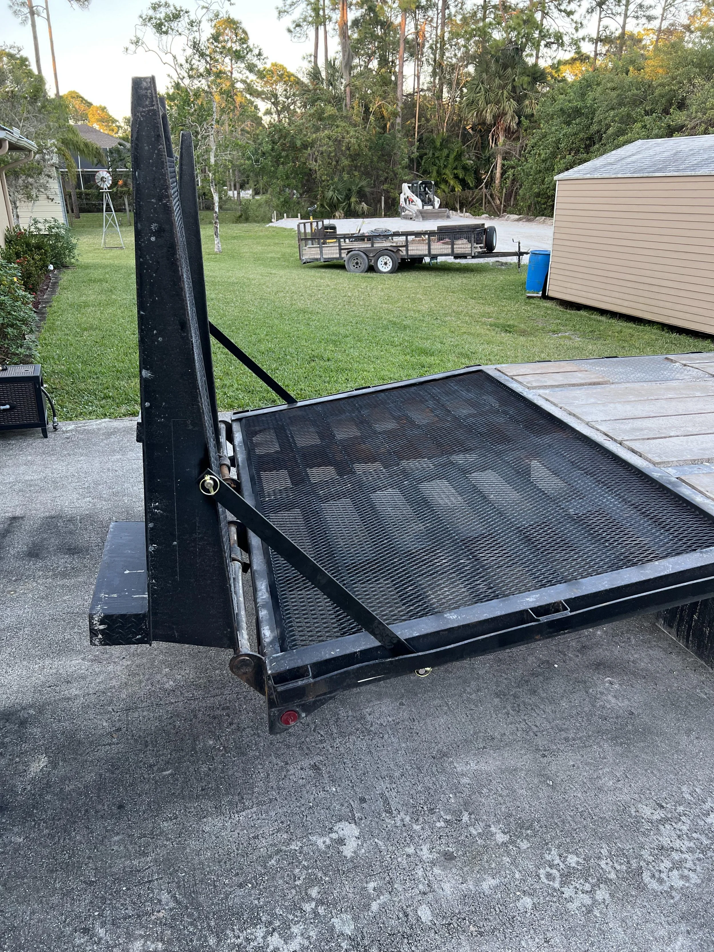 A black flatbed trailer parked on a driveway in a backyard with green grass, trees, a beige shed, and outdoor equipment including a small skid steer loader and a windmill decoration.