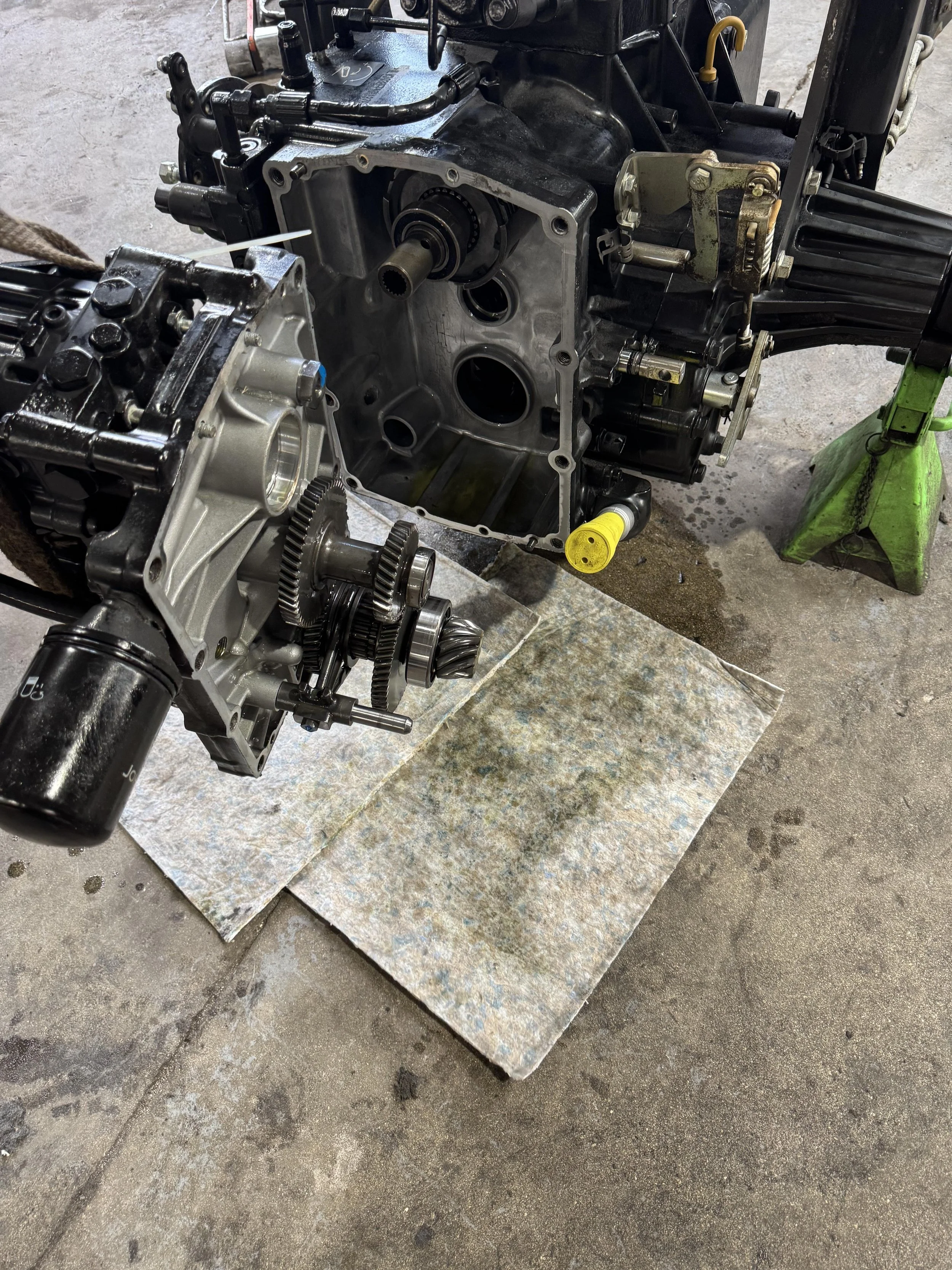 Close-up of a disassembled automotive engine and transmission parts on a workshop floor, with the engine mounted on a stand and the transmission placed nearby on a mat.