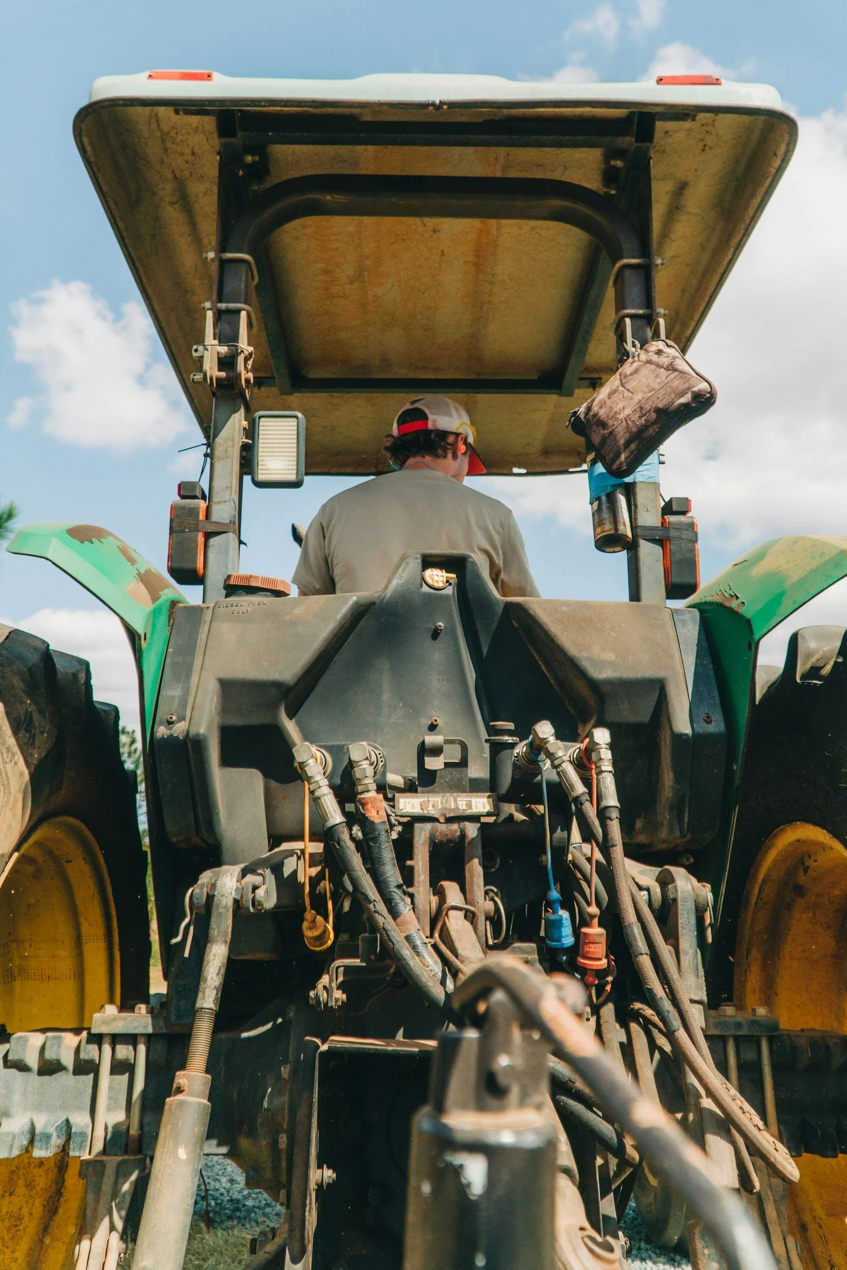 Rear view of a person operating a large green tractor with a yellow wheel, focusing on the tractor's mechanical parts and hydraulic system, under a partly cloudy sky.