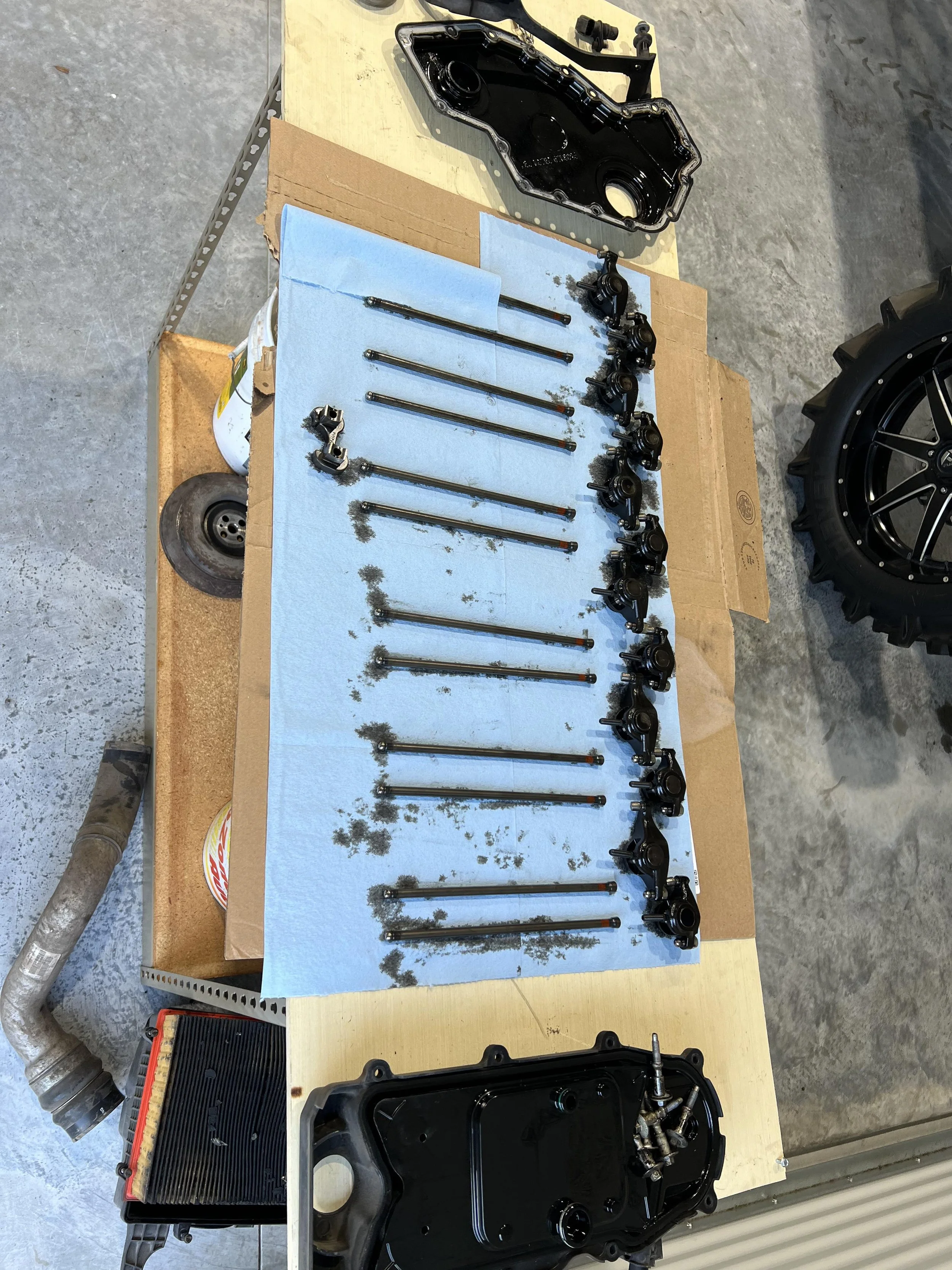 Disassembled automotive engine components laid out on a work table, including valve covers, pushrods, and other engine parts.