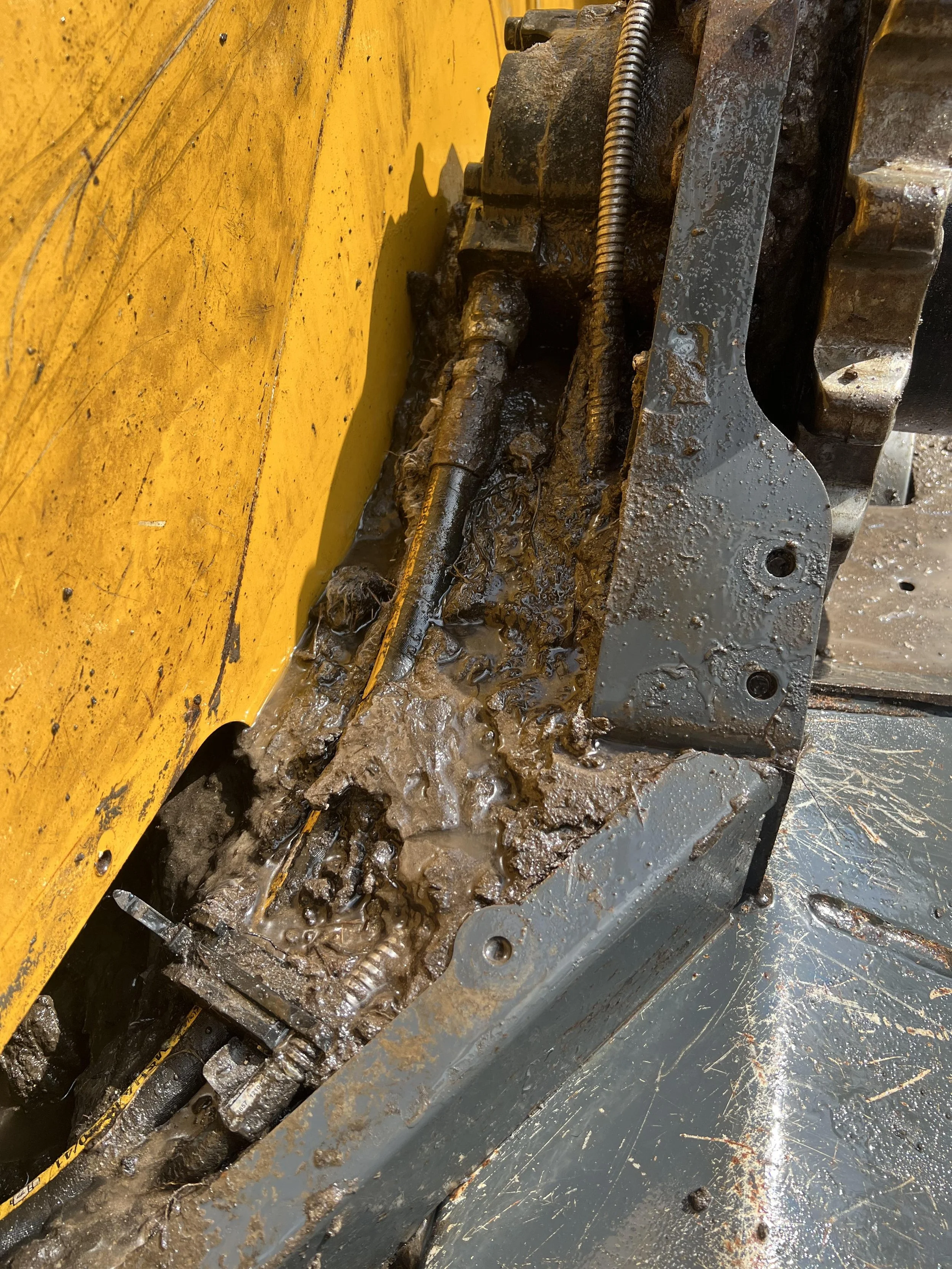 Close-up of a hydraulic system in a construction machine, showing greasy, muddy mechanical parts, metal rods, and a yellow painted surface.