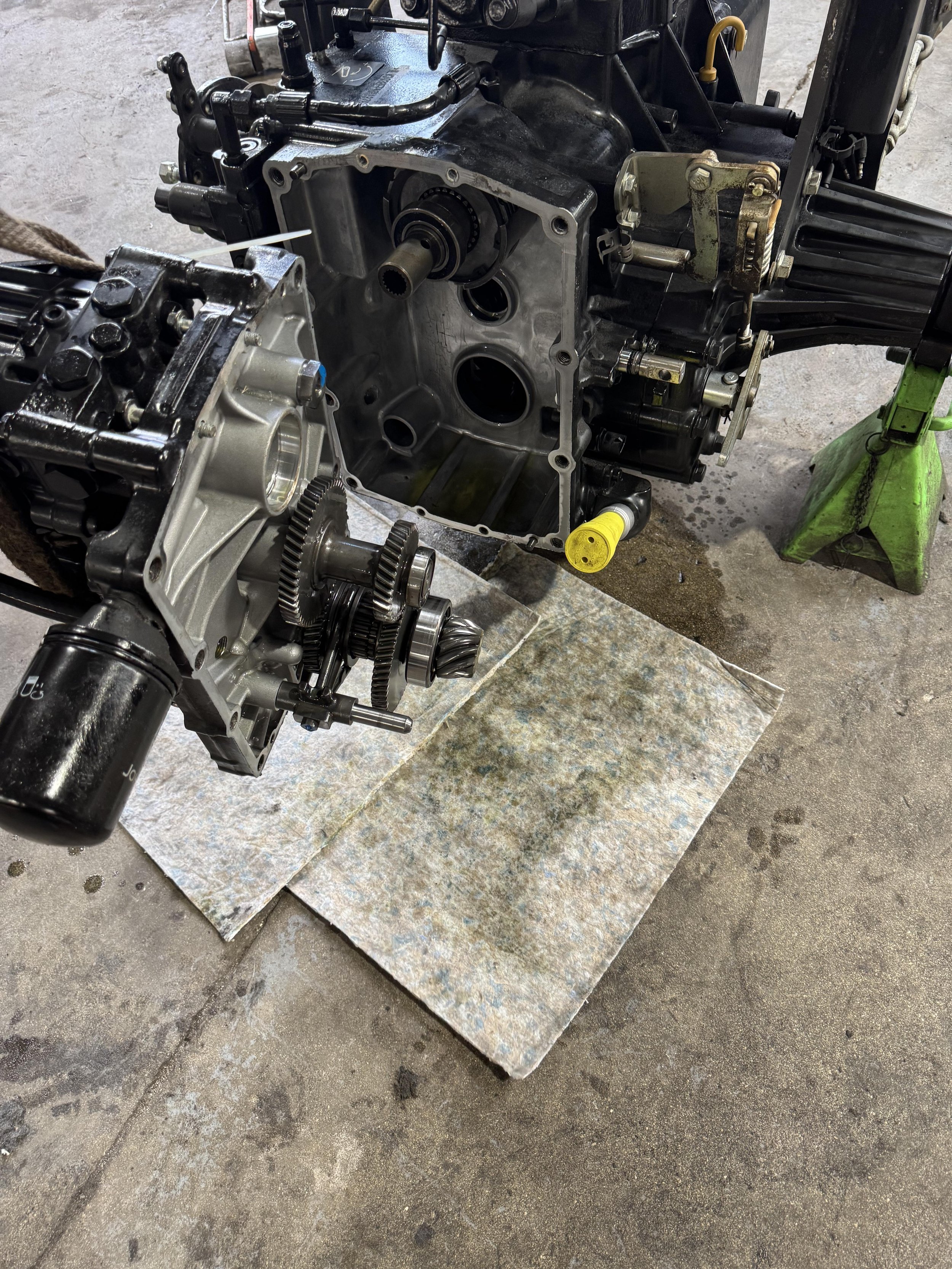 Close-up of a disassembled car engine on a workshop floor with some oil stains, metal sheets underneath, and a green stand supporting part of the engine.