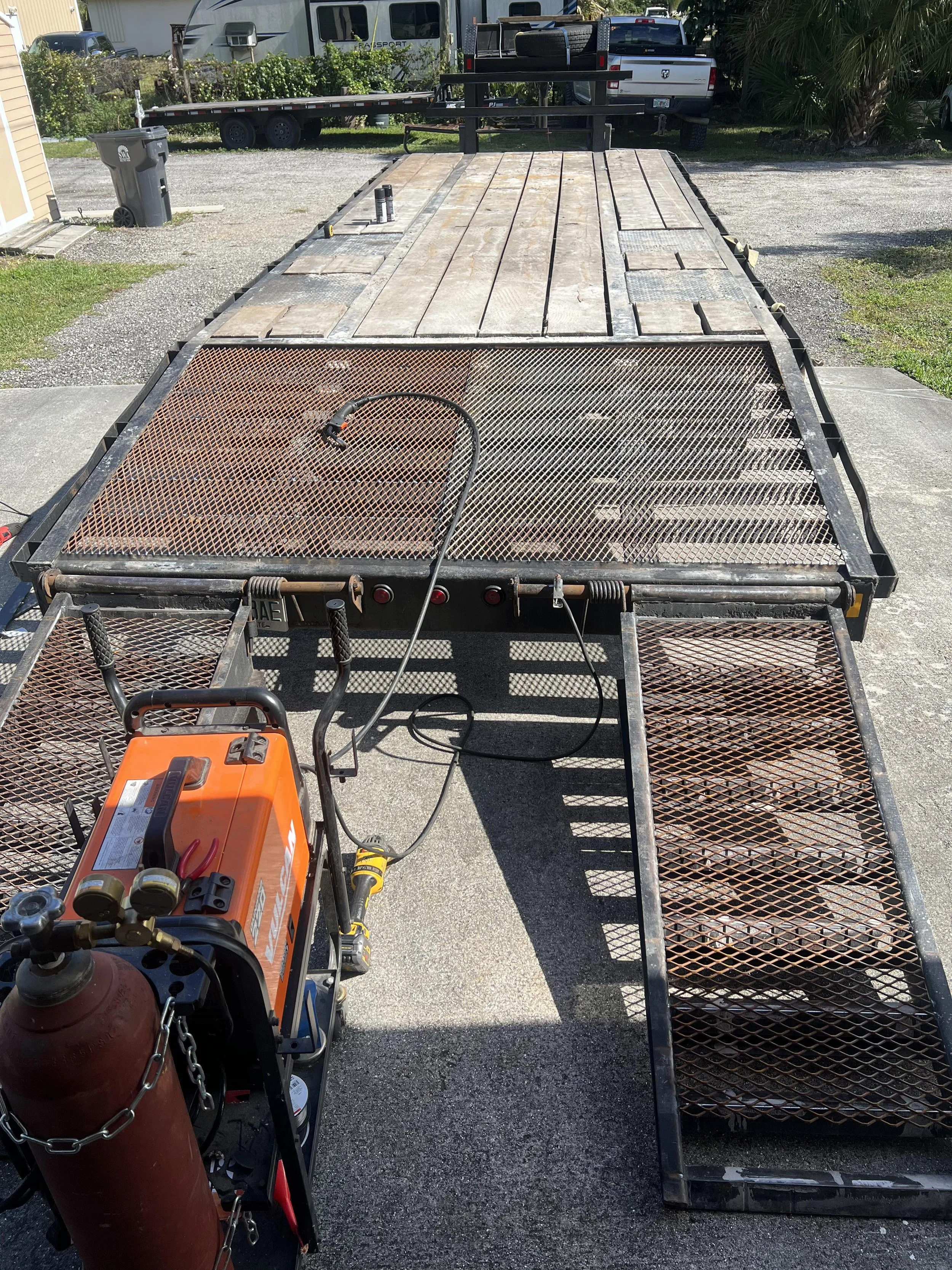 A flatbed trailer with a metal mesh ramp at the back, parked on a paved driveway. The trailer is empty, with some tools and a welding machine placed on it. In the background, there are vehicles, trees, and a storage shed.
