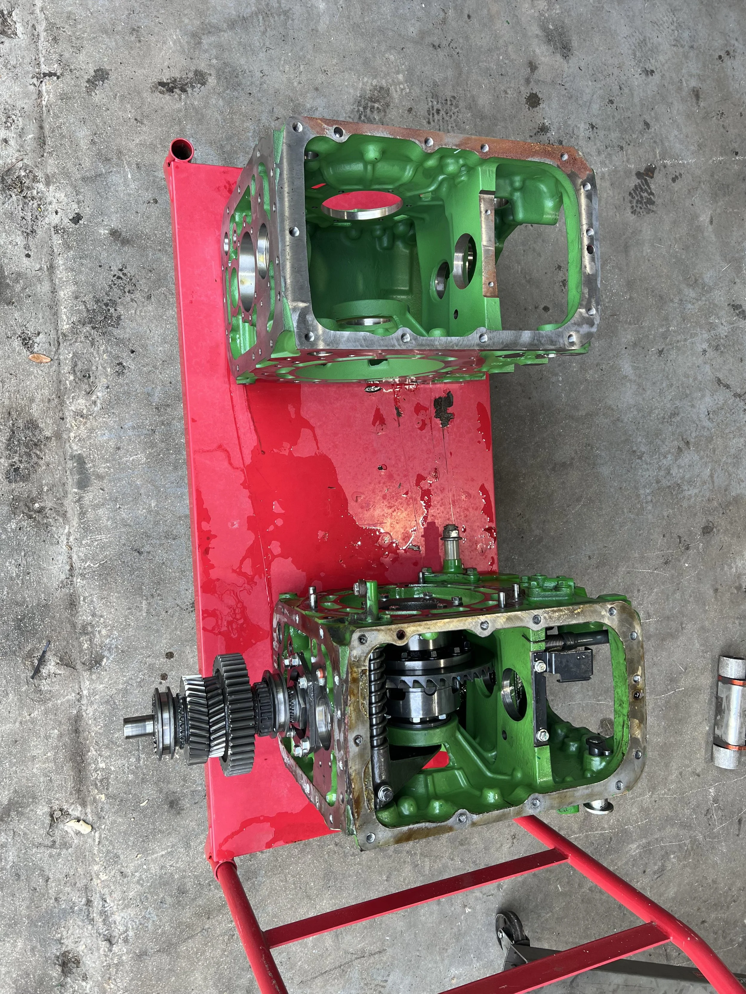 A partially assembled green car engine on a red stand, with gears and mechanical components visible, placed on a concrete floor.
