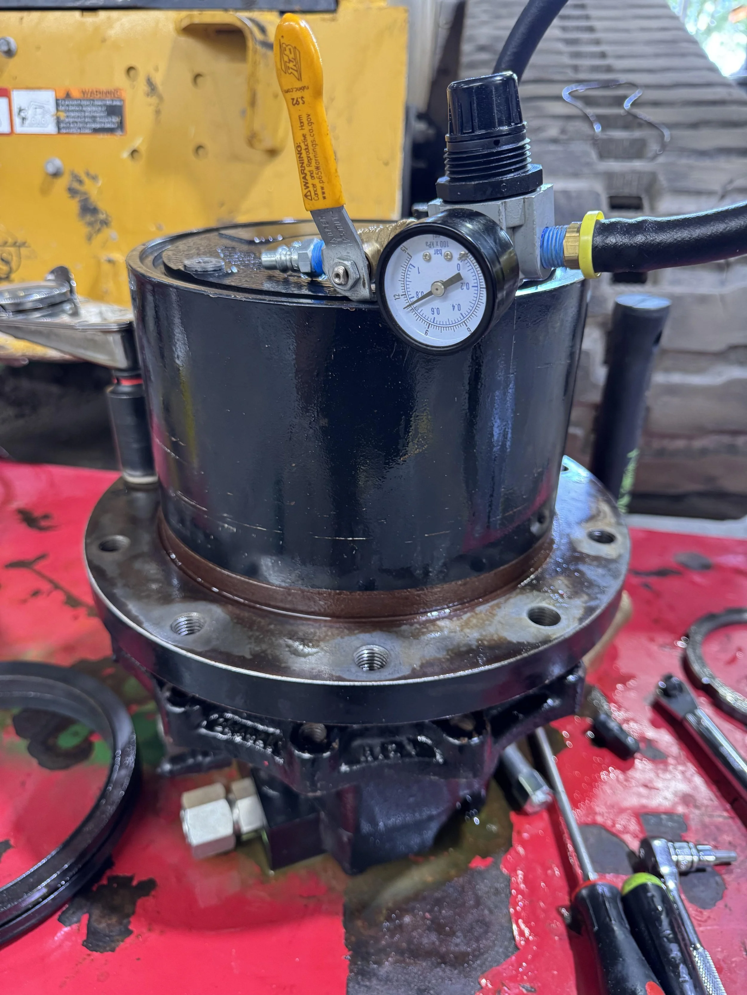 Close-up of a hydraulic pump with a gauge displaying pressure, surrounded by tools on a workbench with red surface, in a workshop setting.