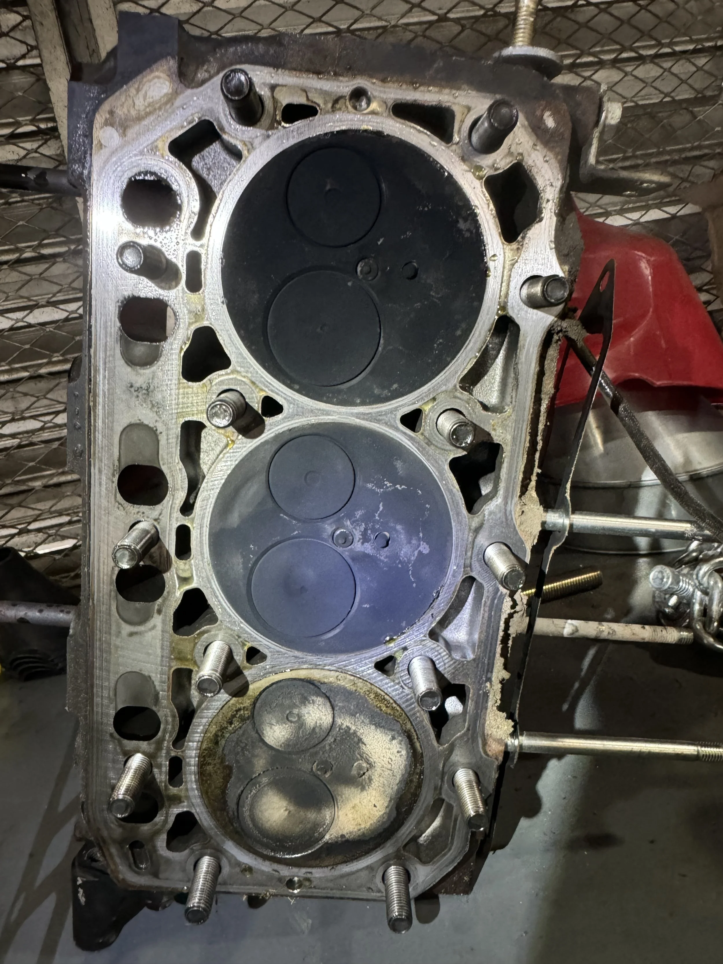 Close-up of a disassembled car engine's cylinder head with pistons visible, mounted on a workbench in a workshop.