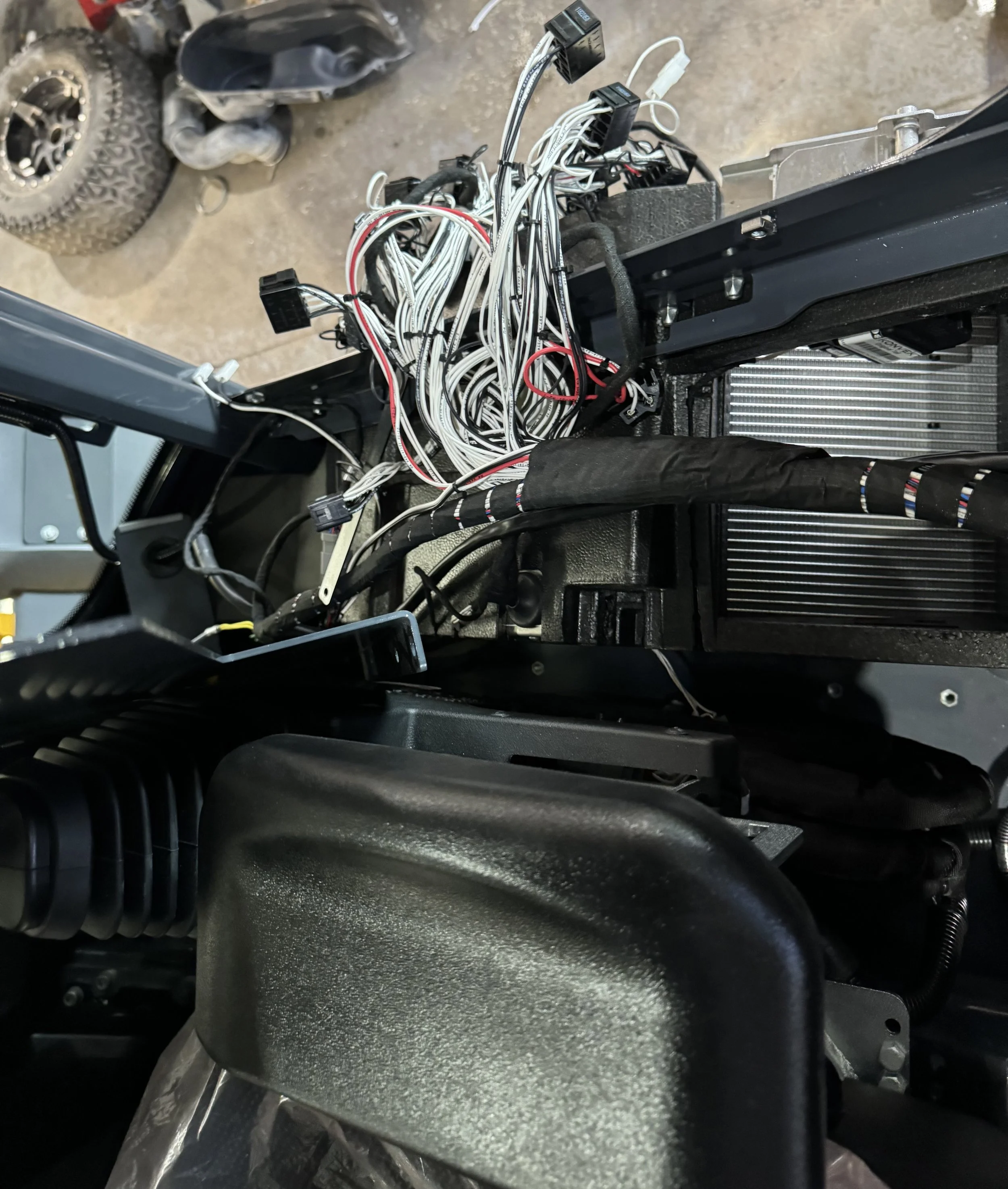 Open computer or electronic device with black and white wires and cables, some protruding and connected to various black modules, next to a metallic radiator or heatsink and other electronic components. The background includes a tire and parts of a w