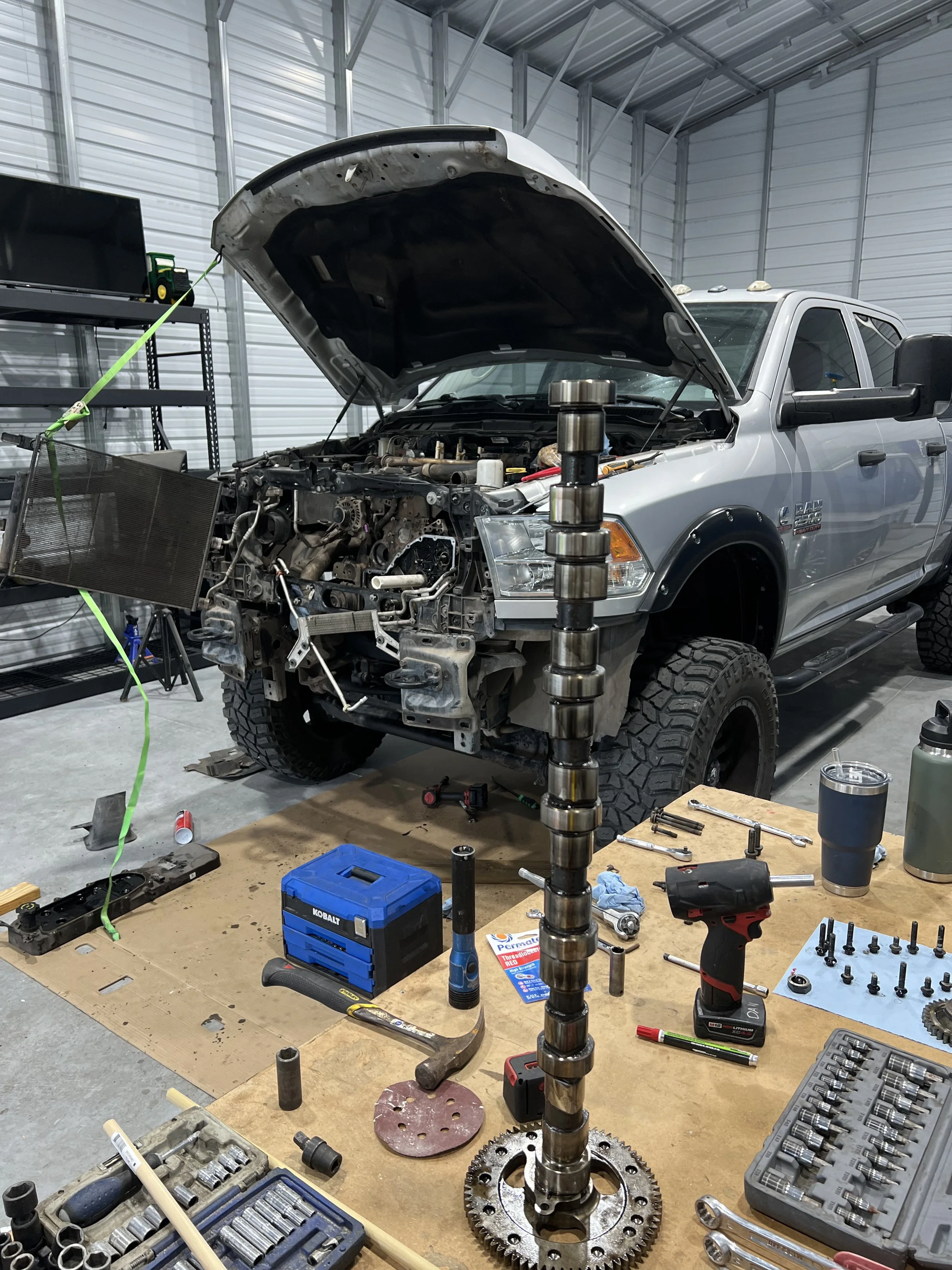 Inside a garage with a pickup truck being repaired, tools, parts, and equipment spread on a workbench and floor, with the truck's hood open and exposed engine parts.
