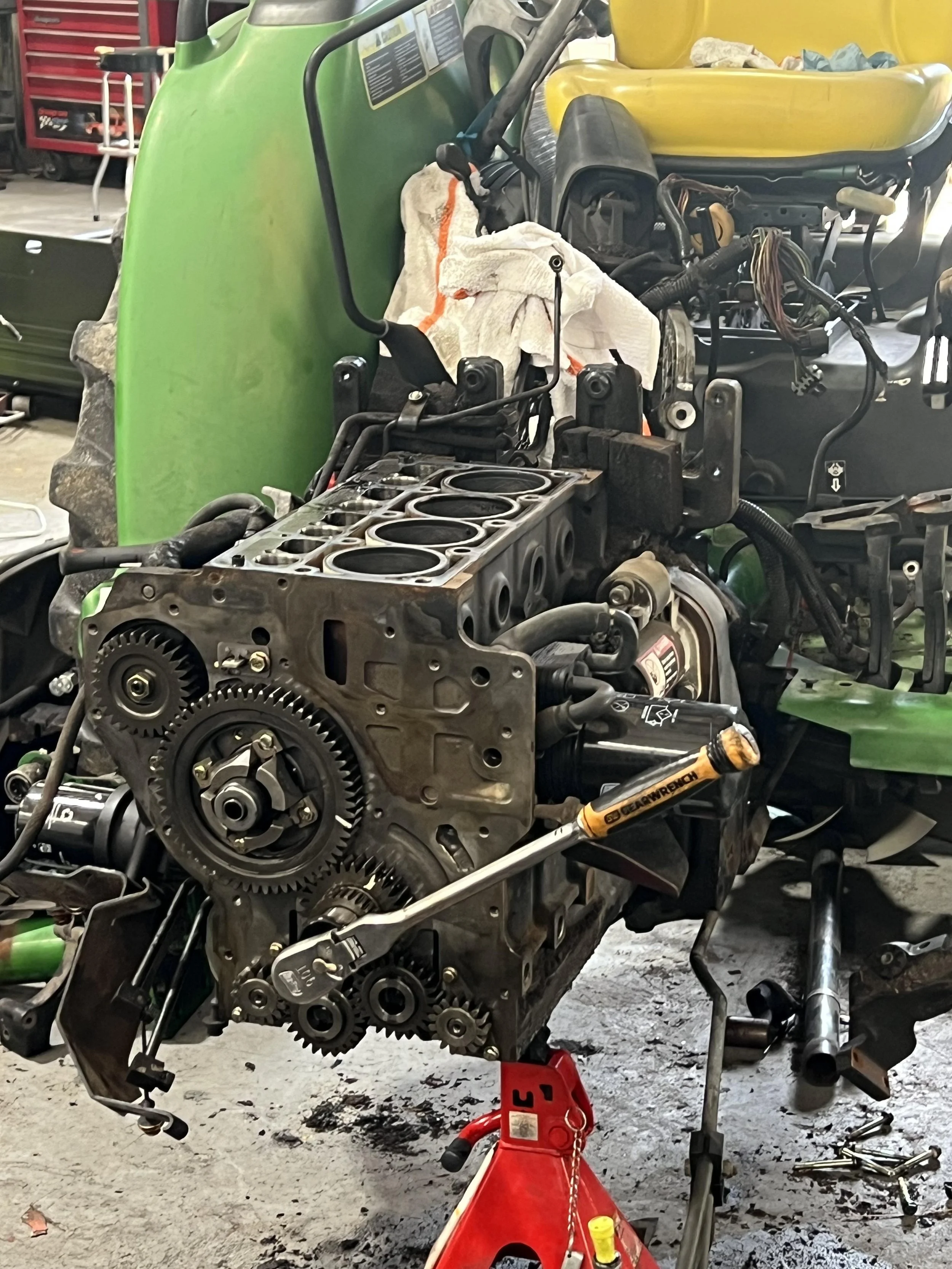 A partially disassembled green vehicle frame with an engine block and various mechanical parts, set in a garage or workshop with tools and equipment in the background.