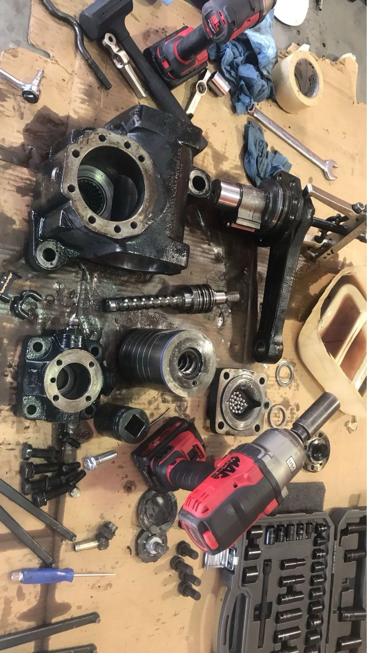 A workbench with disassembled mechanical parts, including a large black metal component, various bearings, bolts, tools, a cordless drill, a socket set in a plastic case, and rags.