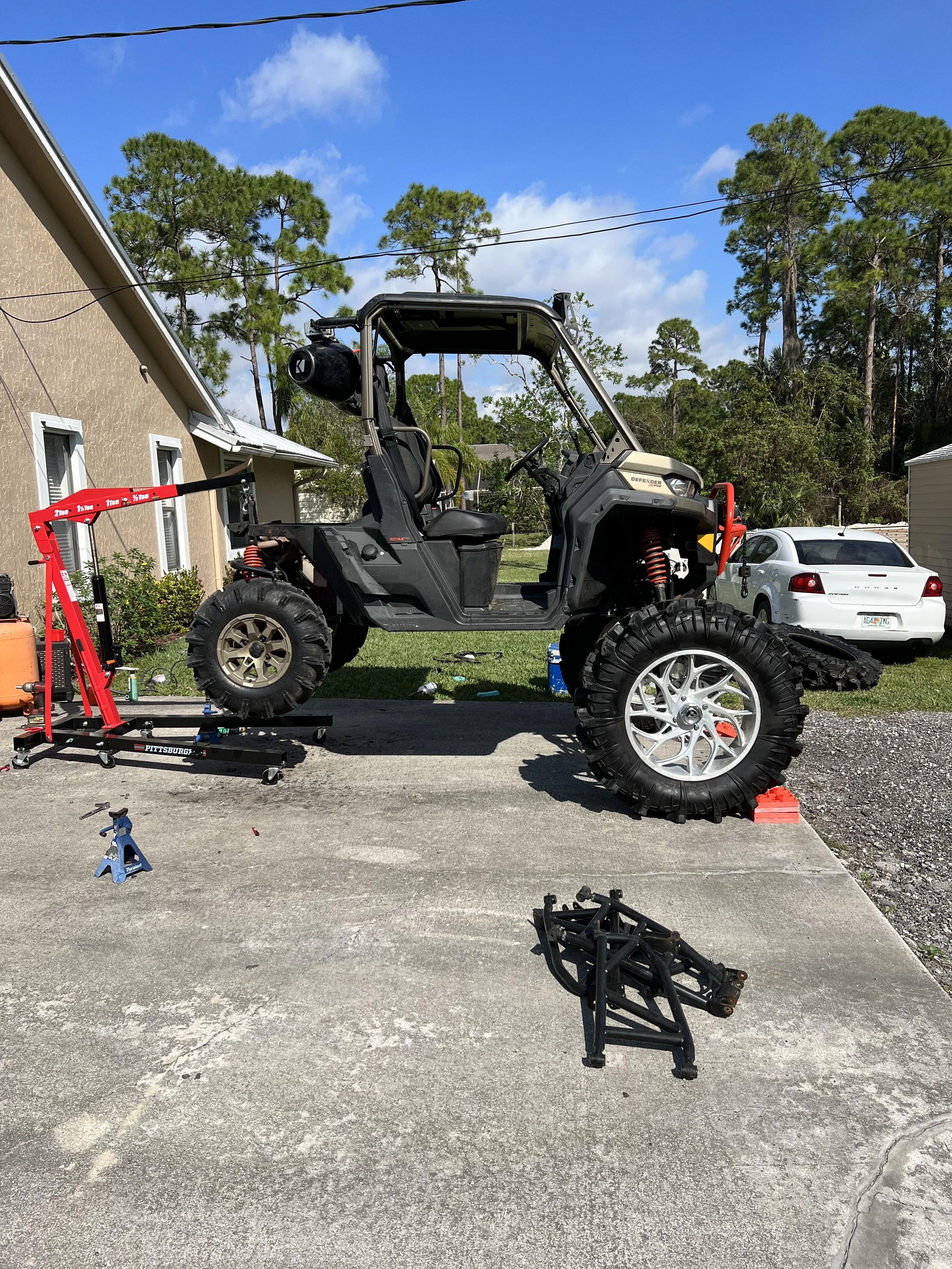 A garage courtyard with a black utility vehicle on a hydraulic lift, a white car parked in the background, and disassembled vehicle parts on the ground. Trees and houses are visible in the background under a blue sky.