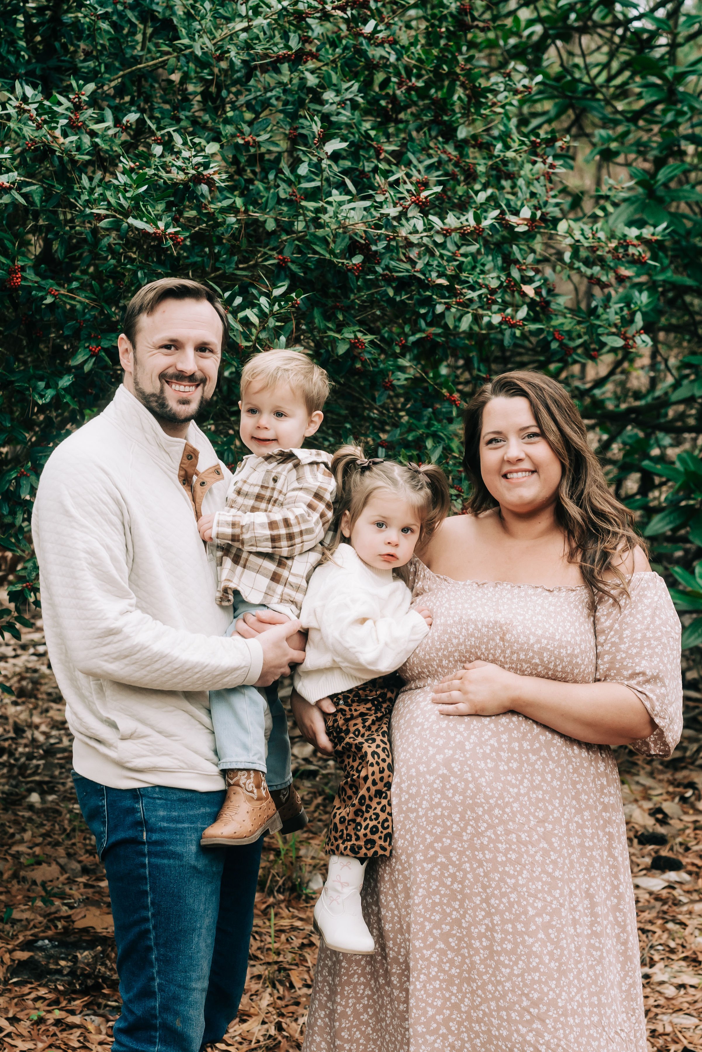A family of four outdoors standing in front of a bush with green leaves and red berries. The father is holding a young boy, and the mother is holding a young girl. The family is smiling and looking at the camera.