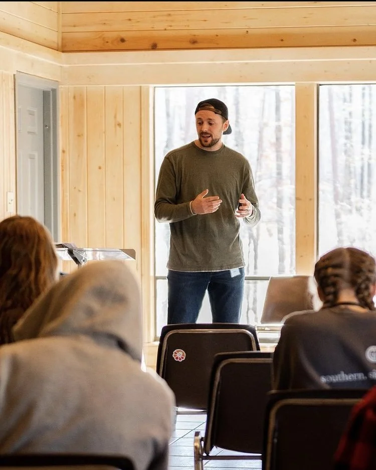 A man giving a presentation to a small group in a room with wooden walls and large windows, with trees visible outside.