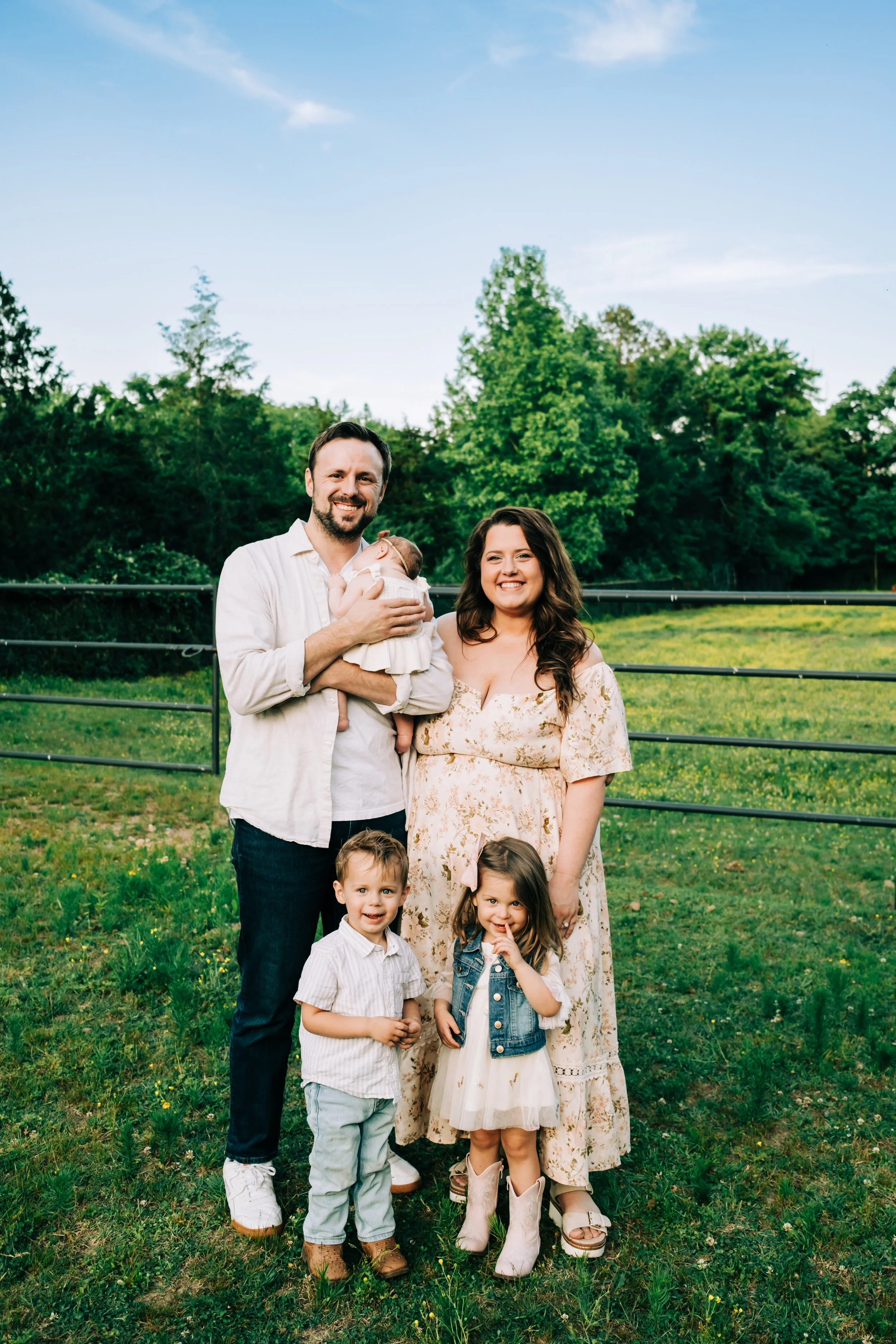 A family of seven outdoors standing on grass in front of a green, wooded area with a blue sky overhead. The adults are smiling, with one holding a baby girl, and four children, aged approximately 2 to 8, standing in front.