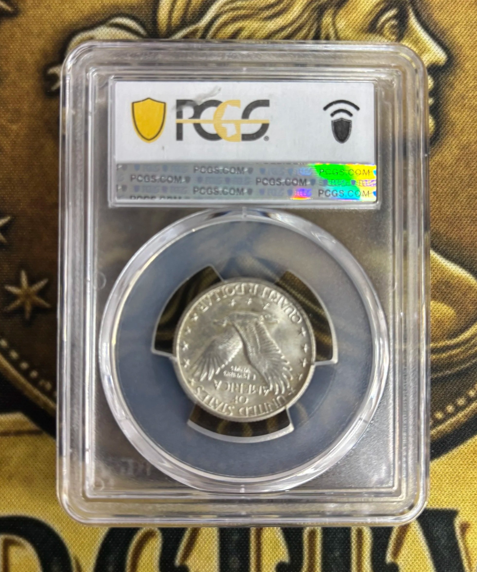 A quarter coin in a protective case with a partial view of the label showing the logo 'PCGS' and a holographic sticker, placed on a fabric with gold and black colors and a lion design.