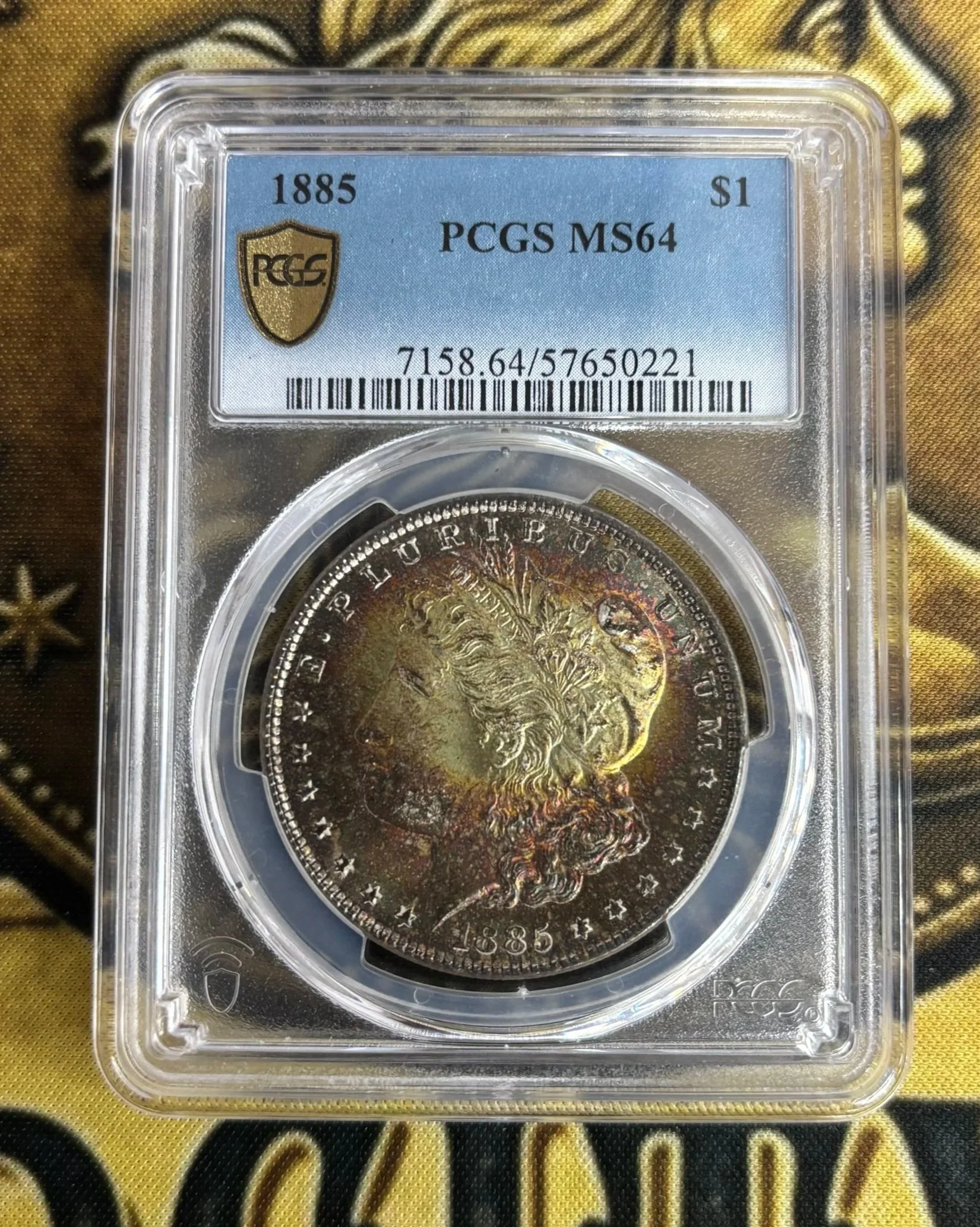A Morgan silver dollar coin from 1885 in protective plastic casing with a label indicating grade MS64, housed on a yellow and black fabric background.