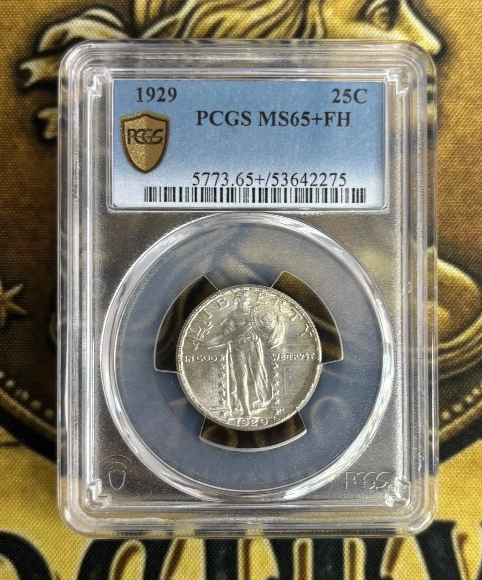 1929 Buffalo Nickel in a plastic holder, graded MS65 by PCGS, with a serial number 5773.65+/53642275, placed on a cloth with a gold and black pattern.