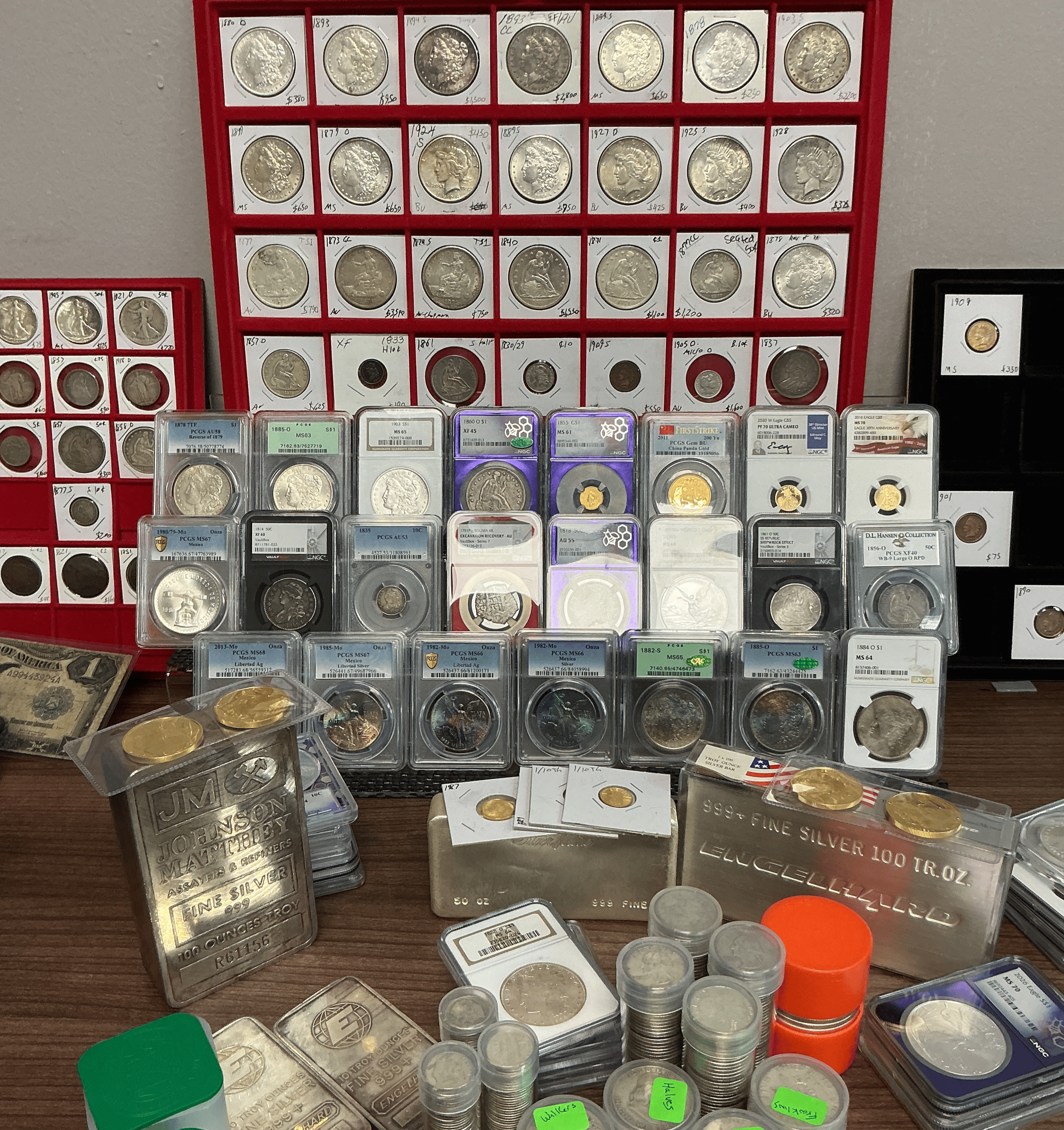 A display of various collectible coins, including silver and gold coins, some in protective cases and others loose. Several dollar bills are also visible, along with orange and green containers, possibly for storage or hobby use. Handwritten price tags and labels are attached to many of the coin cases.