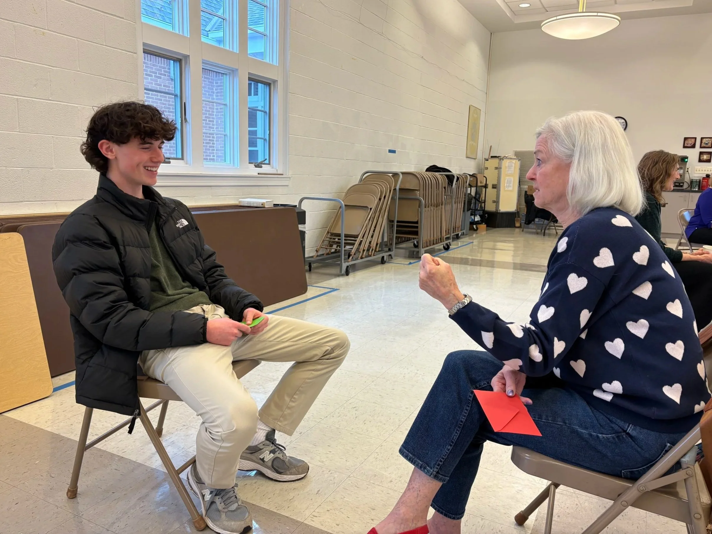 A young man and an elderly woman are sitting across from each other in a room with beige walls and large windows, engaging in a conversation, both smiling. The young man is wearing a black jacket, khaki pants, and gray sneakers, while the elderly woman is wearing a navy sweater with white hearts, a watch, and holding a red card and an orange envelope in her lap.