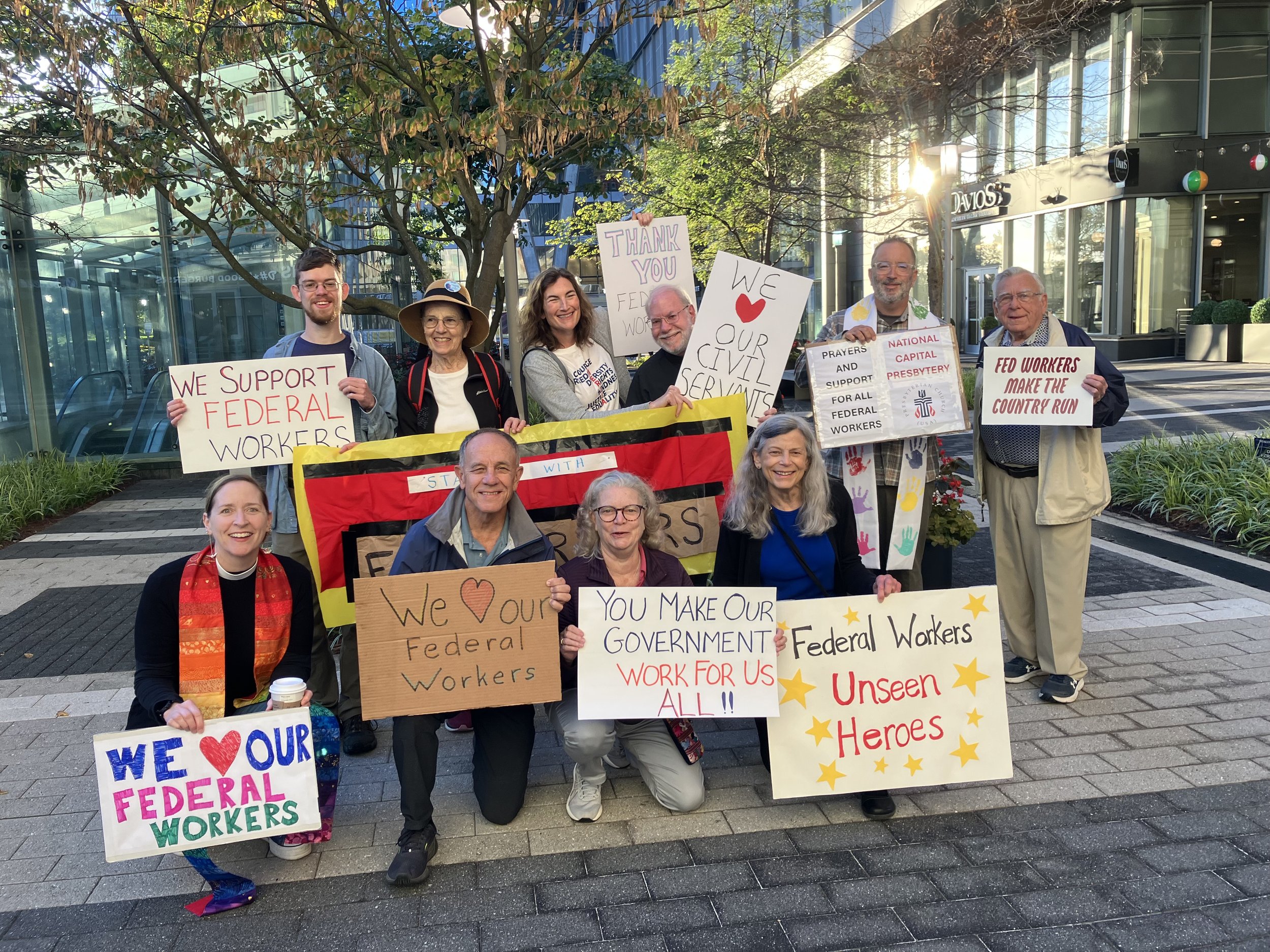 Group of people holding protest signs supporting federal workers and government services outside a modern building with glass windows and a tree. Signs display messages like 'We support federal workers', 'Thank you federal workers', and 'We love our government workers'.