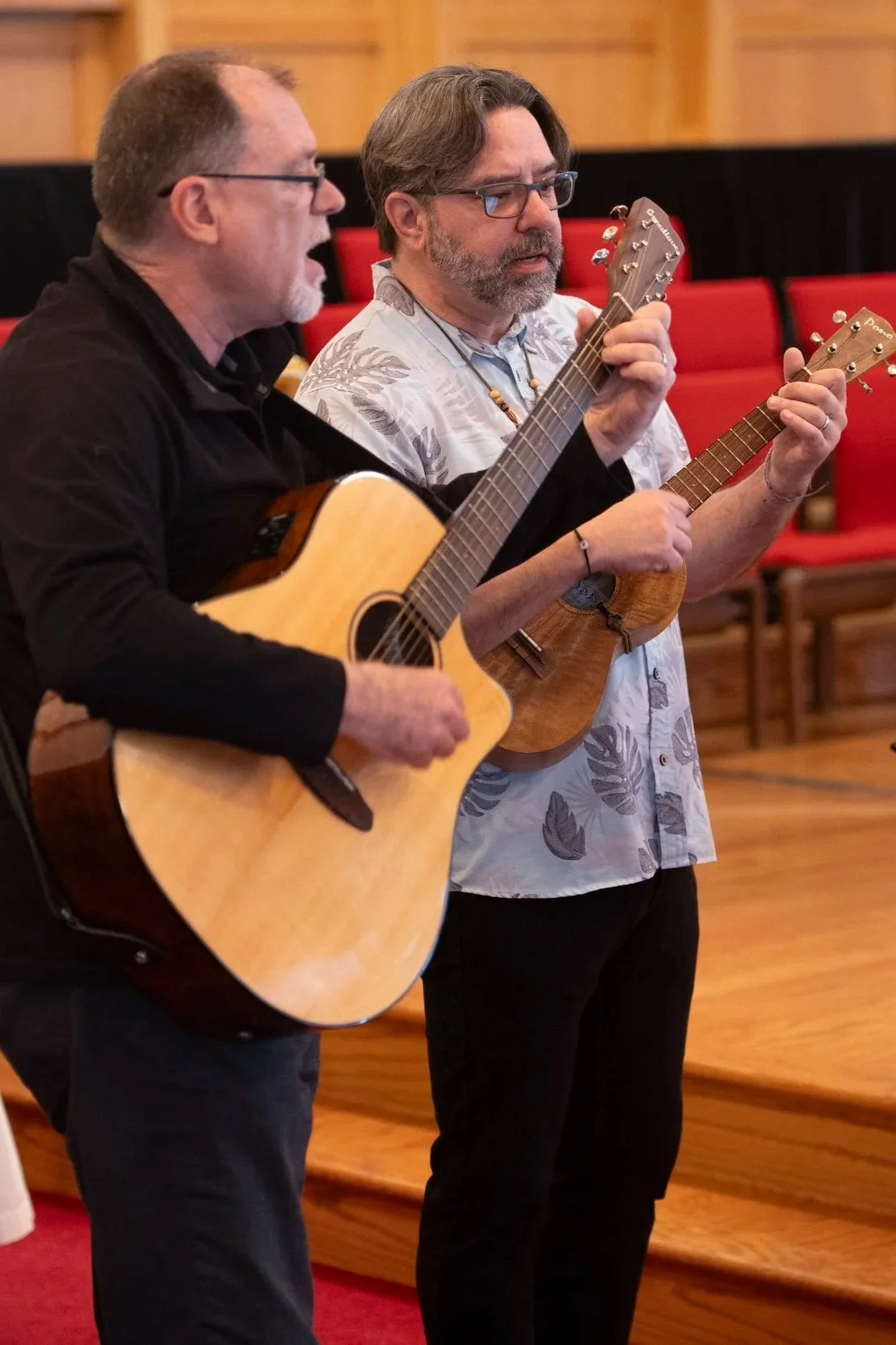 Two men playing guitars, one with an acoustic guitar and the other with a ukulele, in a room with wooden walls and red chairs.