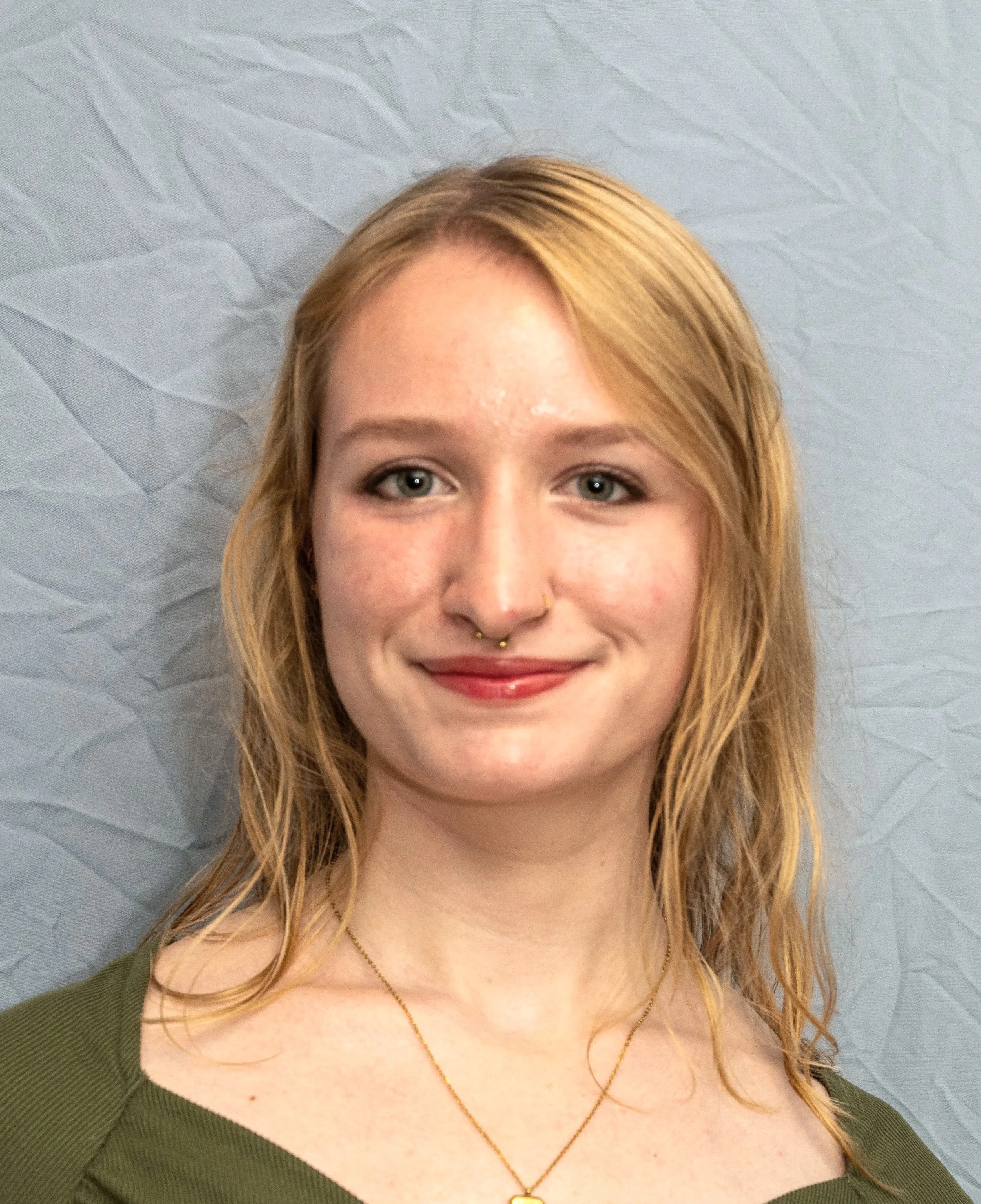 A young woman with blonde hair, blue eyes, and light skin smiling in front blue wall, wearing a green top, gold necklace, and facial piercings.
