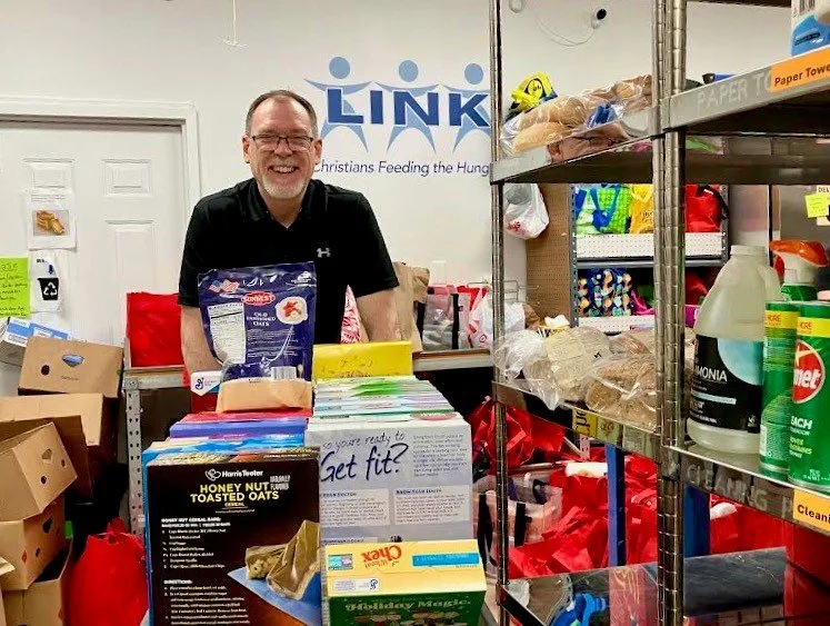 A man in a black shirt standing behind a table with food items in a pantry or storage room. There are shelves with cleaning supplies and food, and a blue 'LINK' sign in the background.