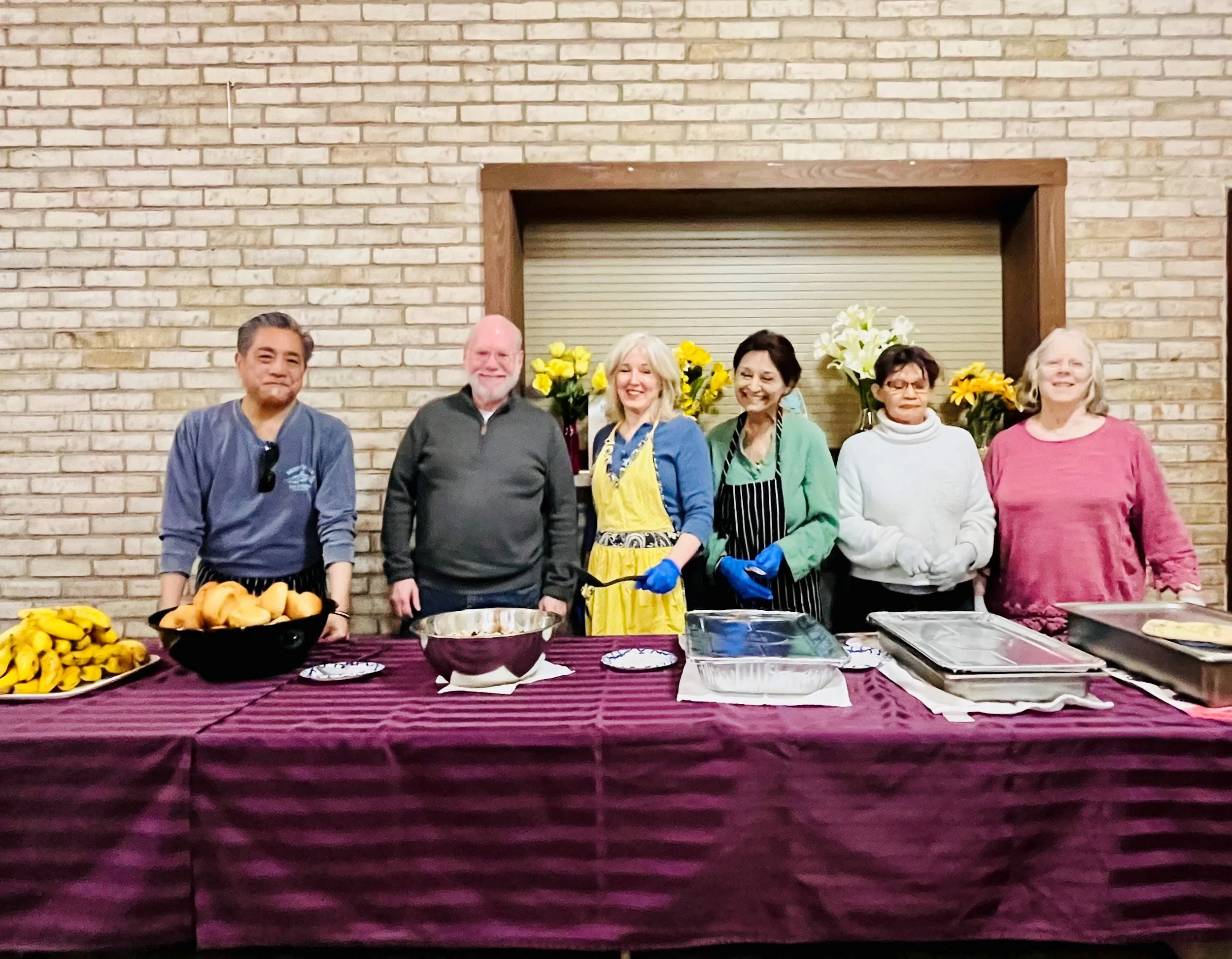 A group of six people standing behind a table with food, flowers, and utensils against a brick wall. They are smiling and wearing casual to semi-formal clothing, with some in aprons and gloves, indicating a gathering or event.