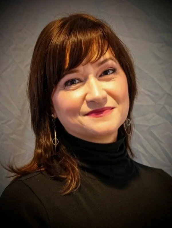 Close-up of a woman with brown hair and earrings, smiling, wearing a black top, in front of a textured white wall.