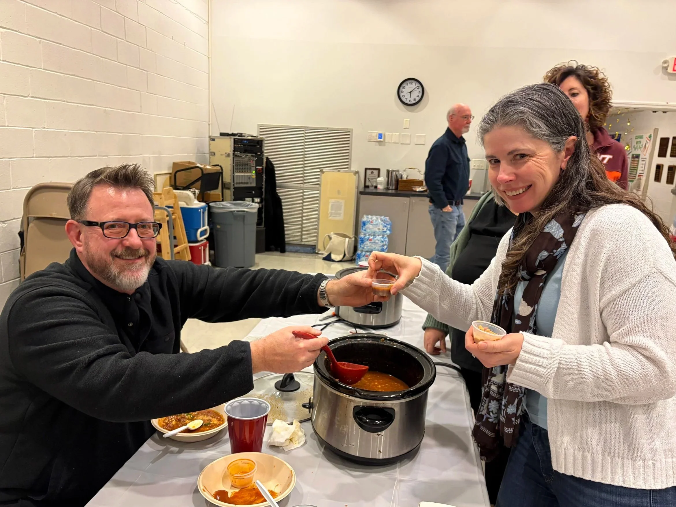 A man with glasses and a woman with long gray hair are smiling and holding hands over a table with a hot pot of soup. There are bowls of food, a red cup, and utensils on the table, and others in the background in a room with a clock on the wall.