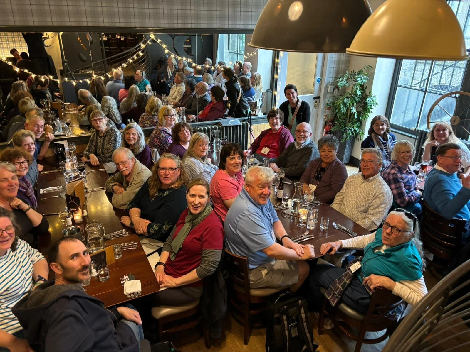 A large group of people gathered around tables in a restaurant, smiling and looking at the camera, with some at the back standing near a railing decorated with string lights, large windows letting in natural light, and greenery.