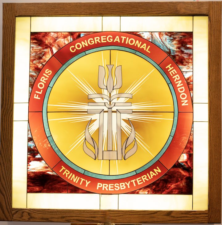 Stained glass window with the Presbyterian Church logo, featuring a cross with flames and a book, surrounded by the words 'Floris Congregational Herndon Trinity Presbyterian' on a red circular band.
