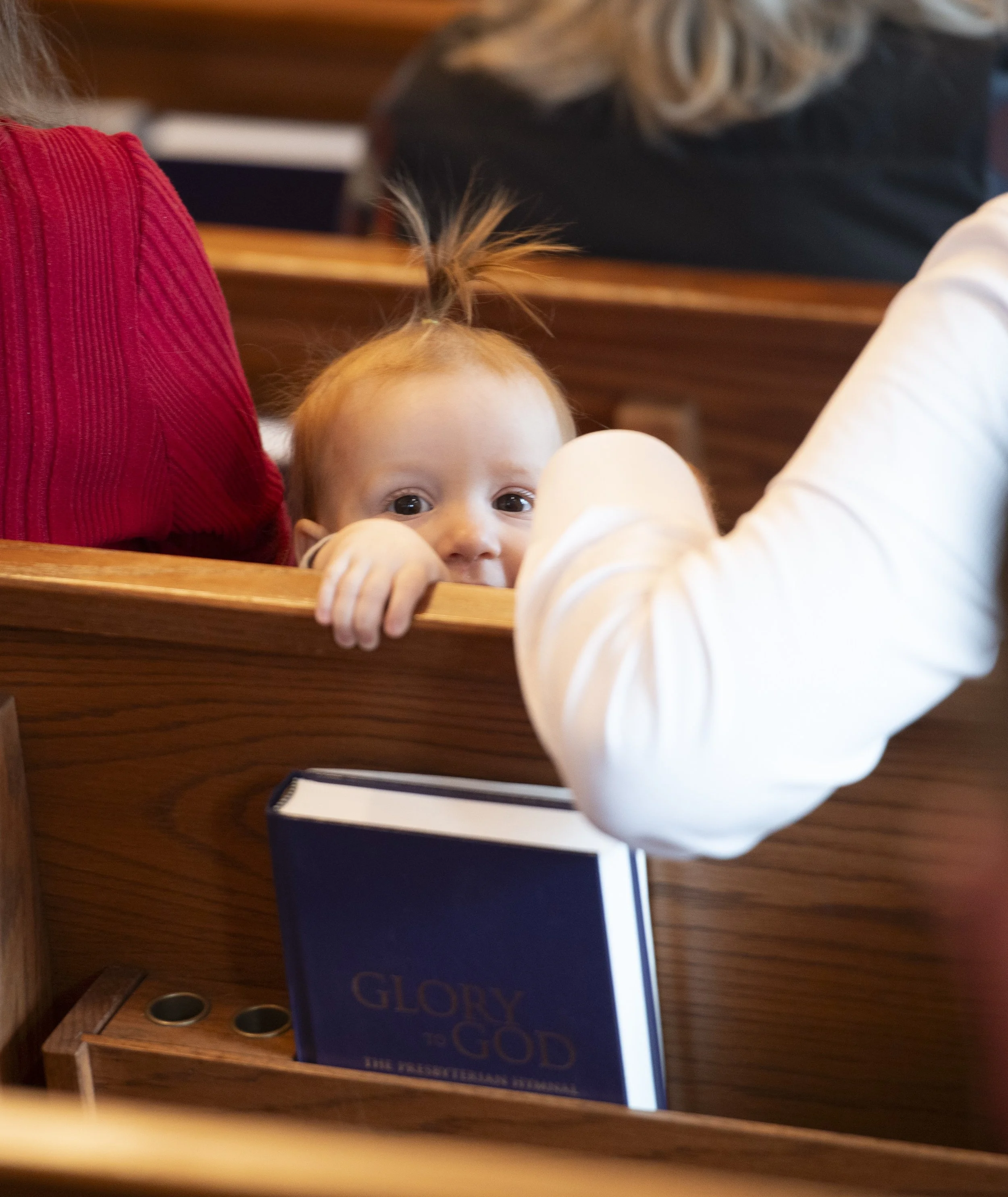 A young child with bright eyes peeking over the edge of a table, with her hands gripping the front of the table, in front of a blue book titled 'Glory to God' on a wooden surface.