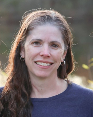 A woman with long wavy brown hair, smiling outdoors with a blurred natural background, wearing a navy blue top and hoop earrings.