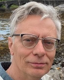 Close-up of a middle-aged man with gray hair and glasses, outdoors near a body of water with rocks in the background.