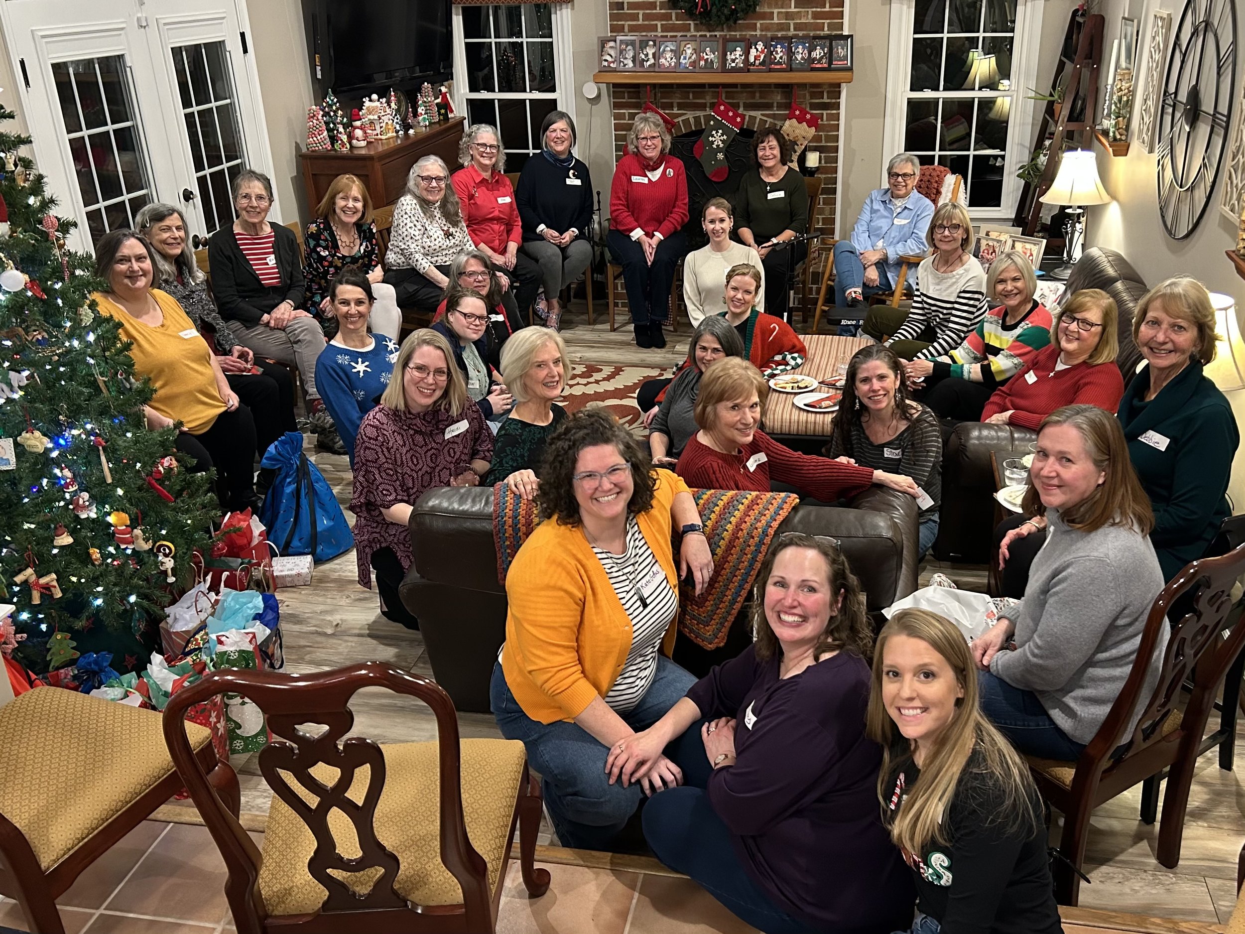 Group of women gathered in a living room decorated for Christmas, with a Christmas tree, presents, and holiday decorations including stockings and framed photos on the fireplace mantel.