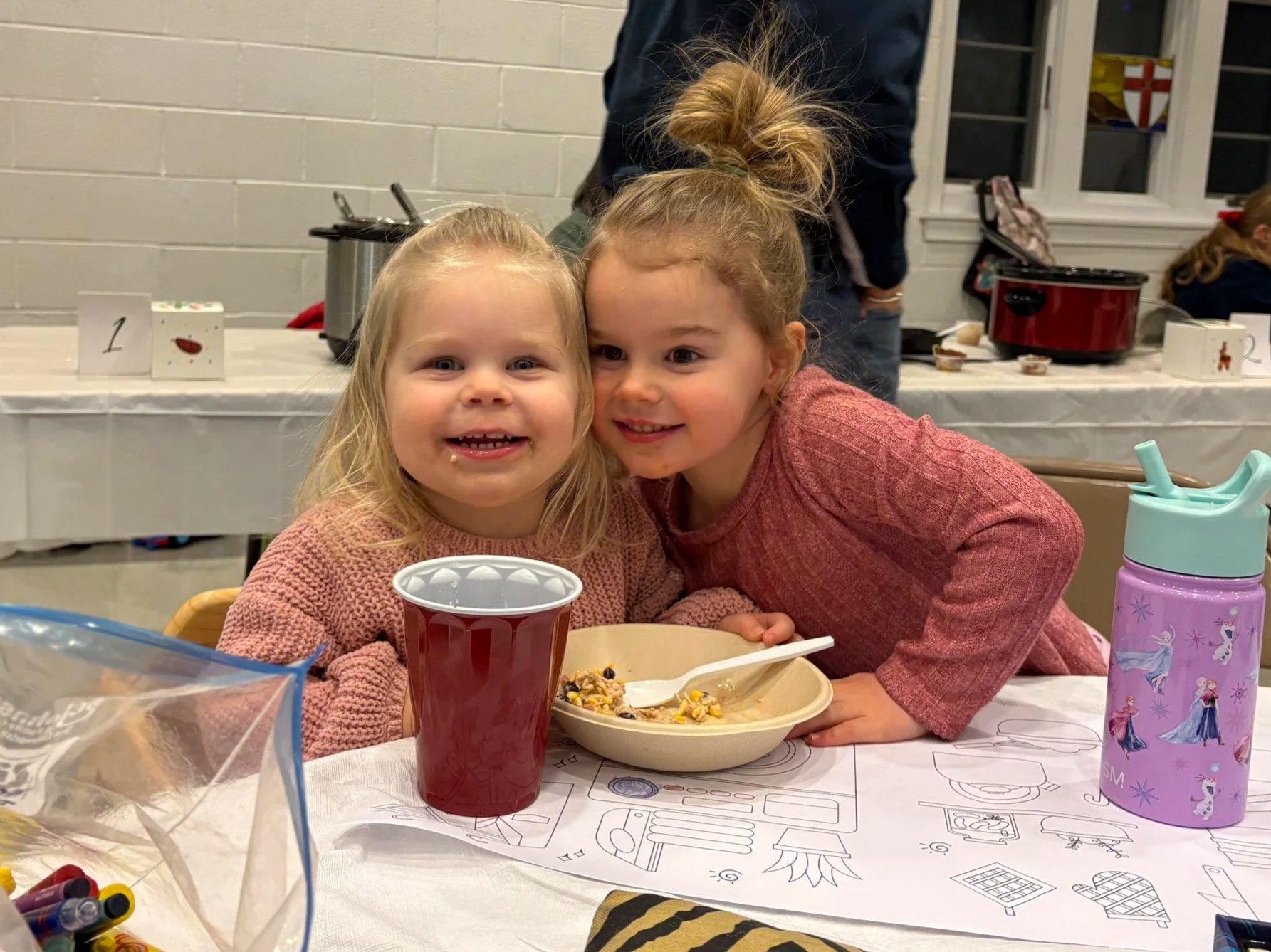 Two young girls sitting at a table, smiling at the camera, with a table full of food and drinks in front of them. The girl on the left has blonde hair and is holding a red cup, while the girl on the right has light brown hair tied up in a messy bun and is wearing a pink sweater. There are coloring sheets and markers on the table, with a water bottle and a pink cup with cartoon characters beside them. In the background, there is a white brick wall, a window, and other children and adults standing, with a dessert table and some kitchen equipment.