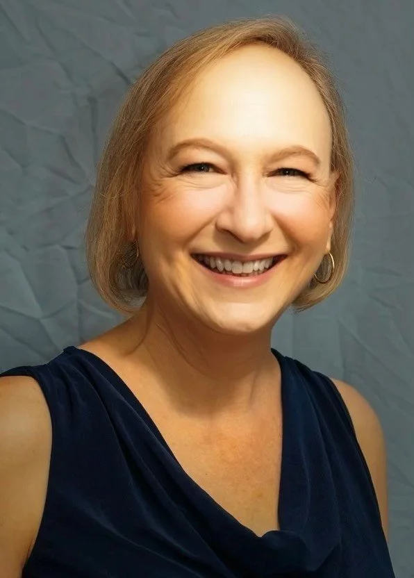 A woman with short gray hair, wearing a dark blue sleeveless dress, smiling, and standing in front of a crinkled blue paper background.