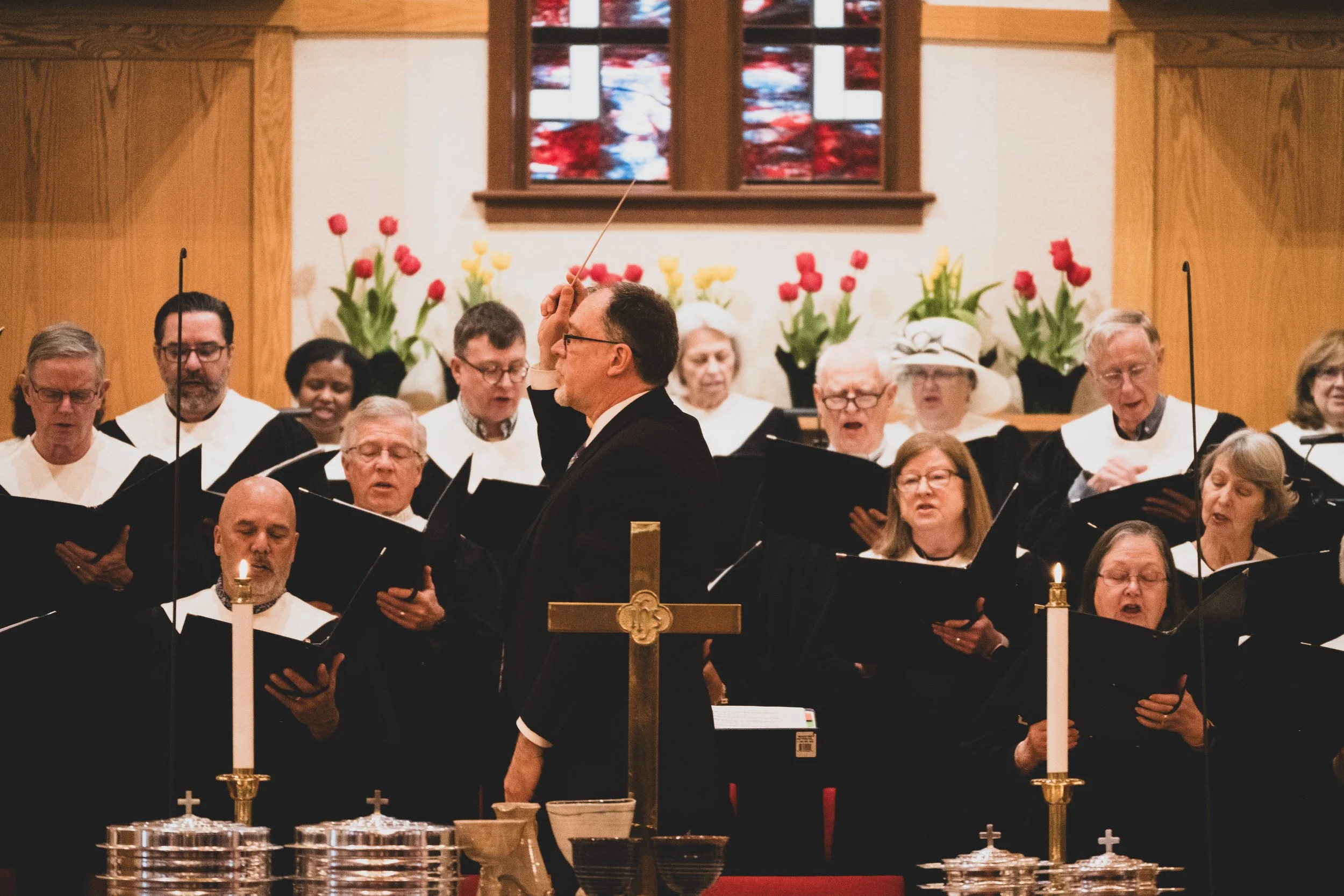 A choir of people singing during church service with a conductor leading, church altar with candles, and floral decorations in the background.