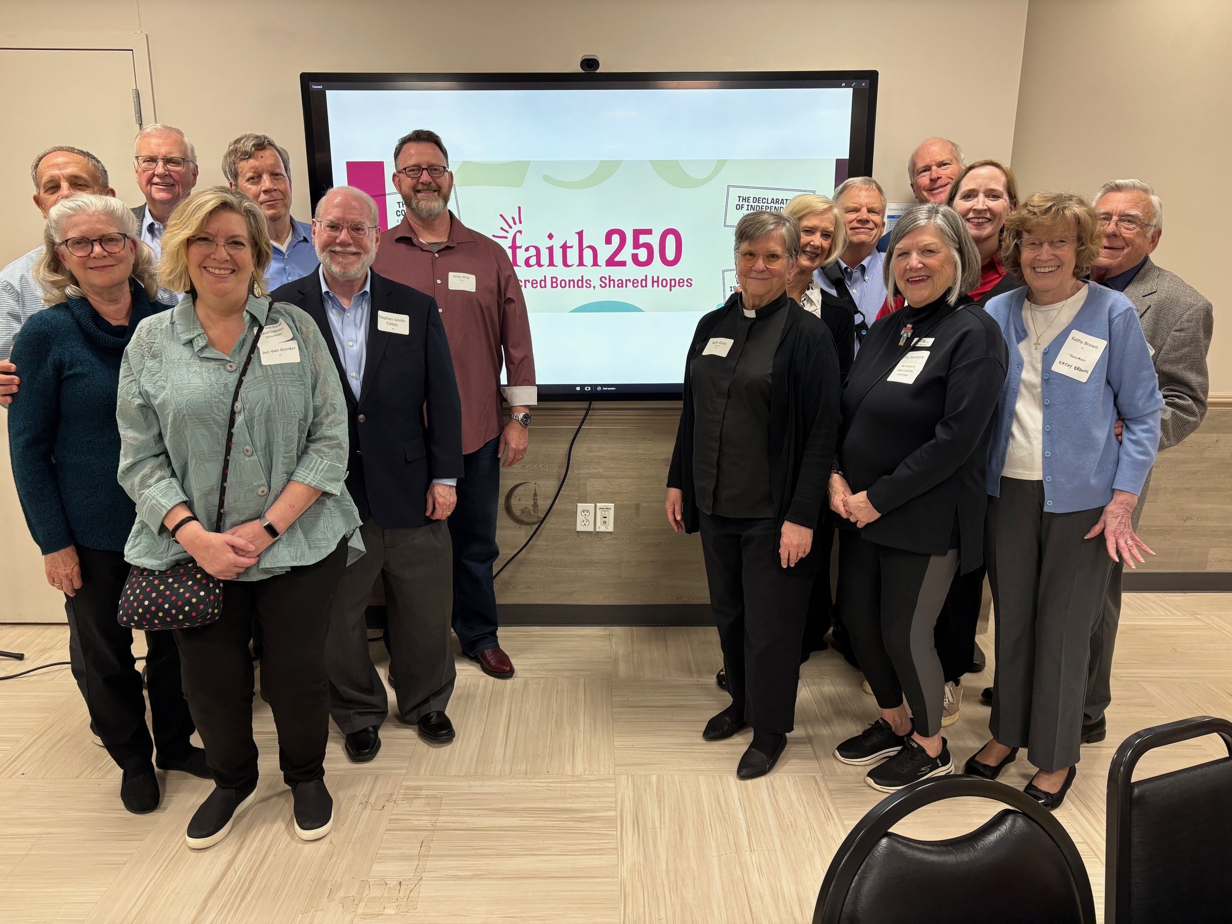 Group of adults standing indoors in front of a large digital screen displaying the words "faith 250" and the phrase "Shared Bonds, Shared Hopes". The group is smiling and wearing casual to business attire.