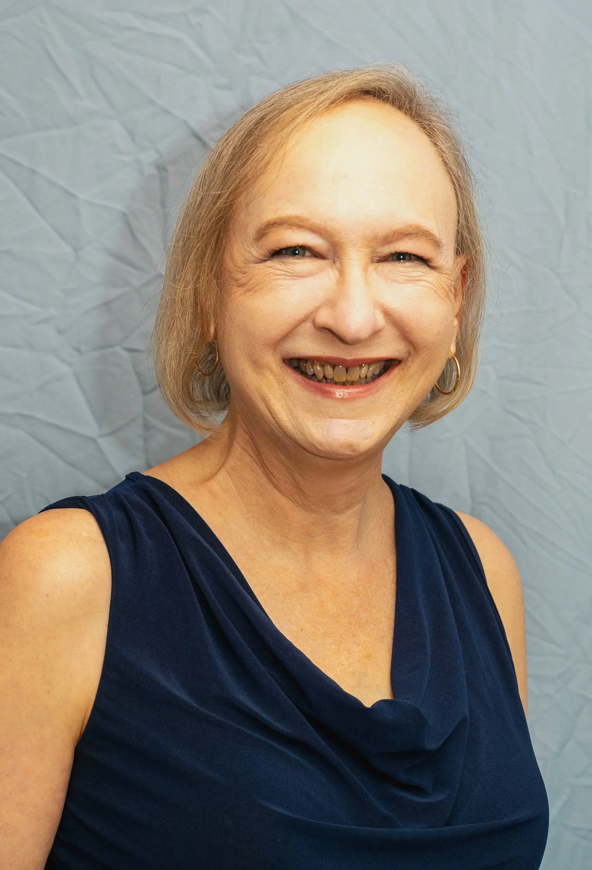 A woman with short gray hair, wearing a dark blue sleeveless dress, smiling, and standing in front of a crinkled blue paper background.