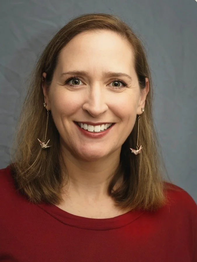 A woman with shoulder-length light brown hair and green eyes smiling, wearing earrings with origami cranes and a red top, standing in front of a plain light gray background.