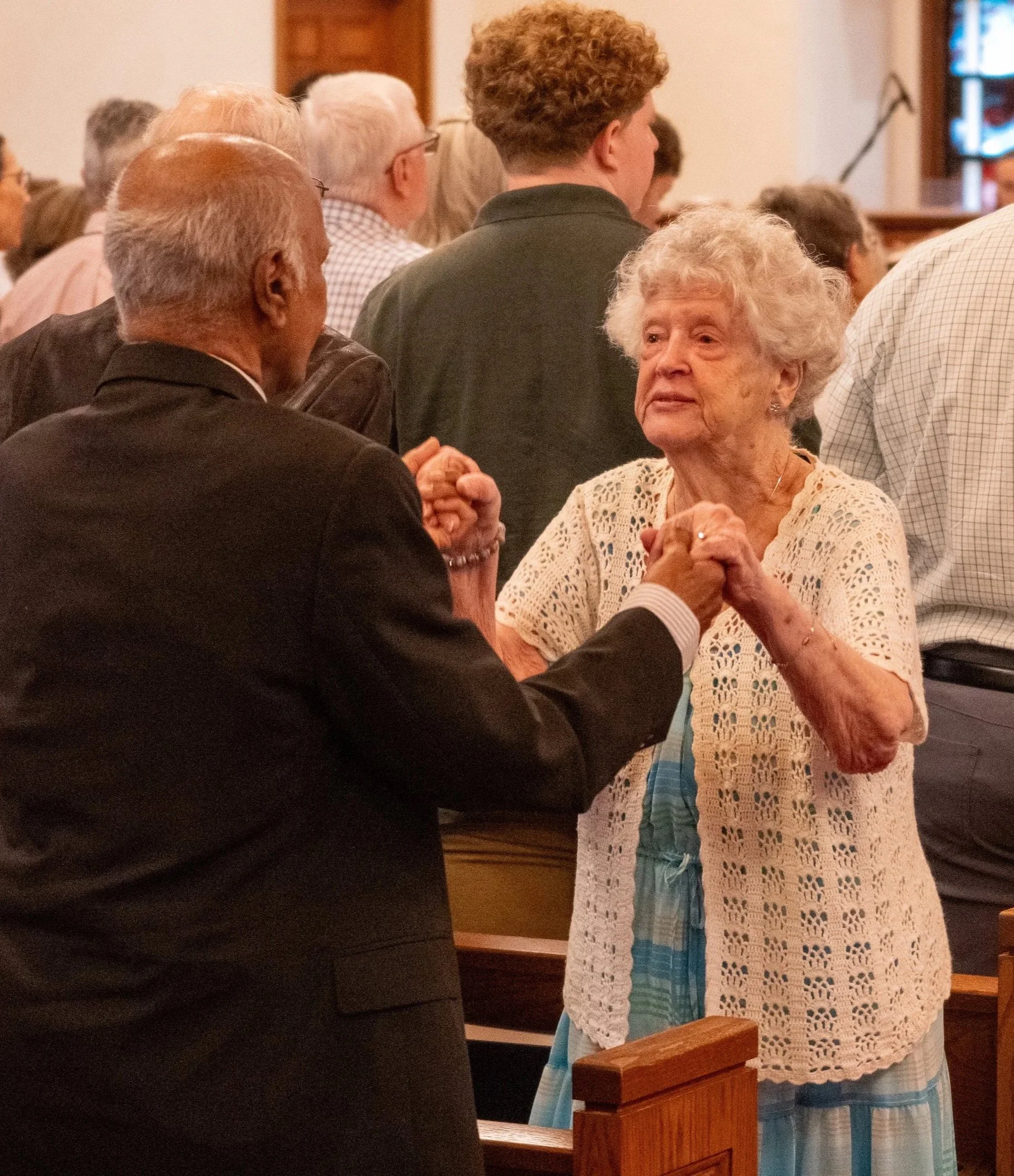 Elderly woman and younger woman holding hands during a church or religious service, with seated congregation in the background.