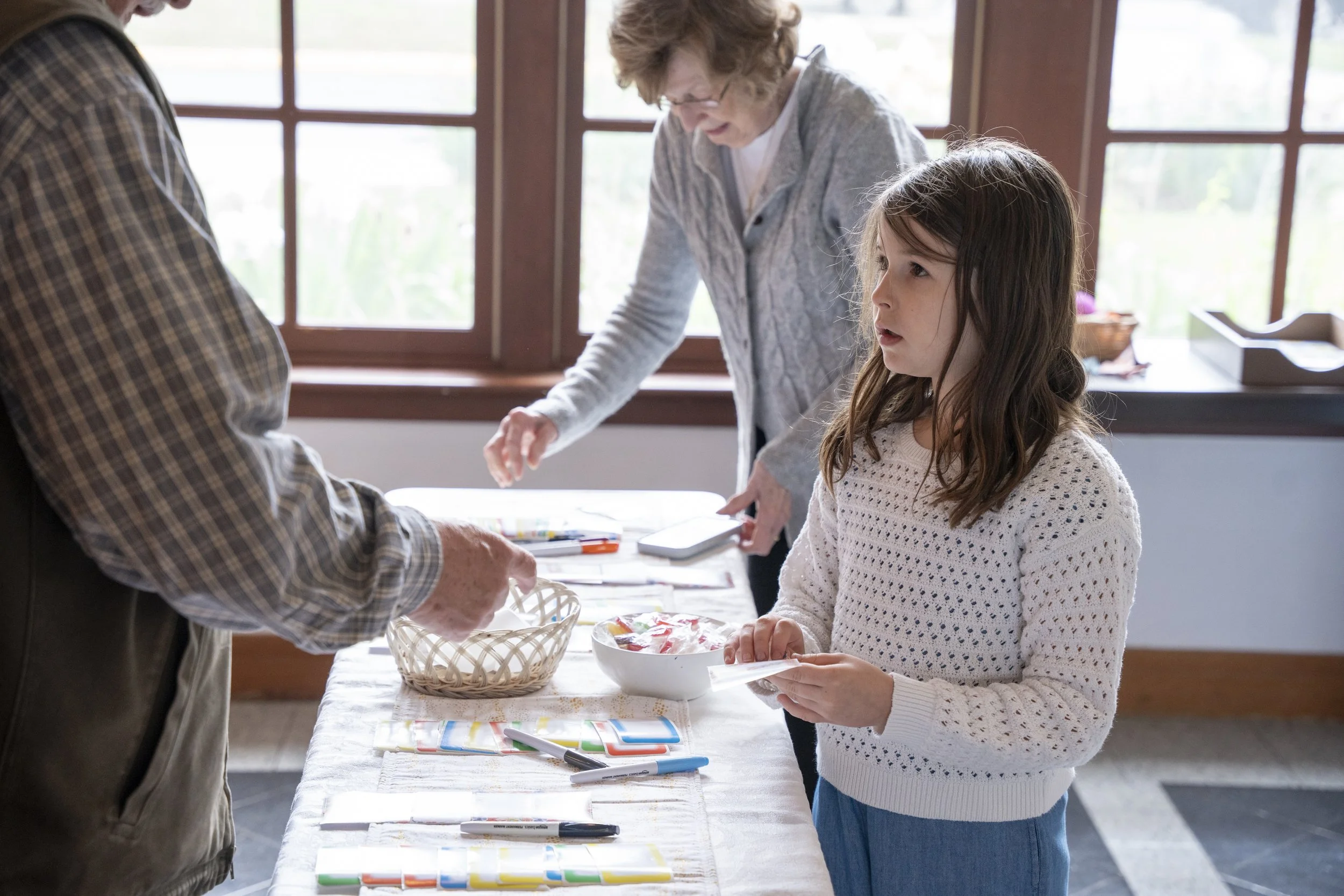 A young girl stands at a table with art supplies, looking attentively while an elderly woman behind her points to something on the table during an activity or craft session.