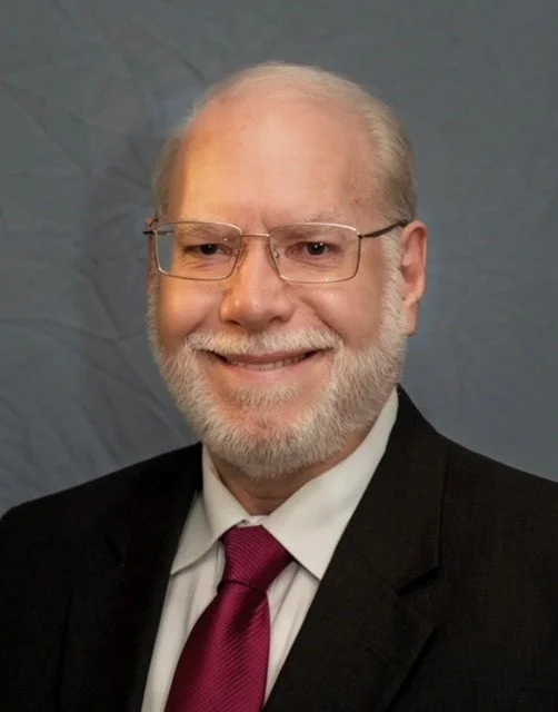 Close-up of an older man with glasses, white hair, and a beard wearing a black suit, white shirt, and magenta tie, smiling in front of a gray patterned background.