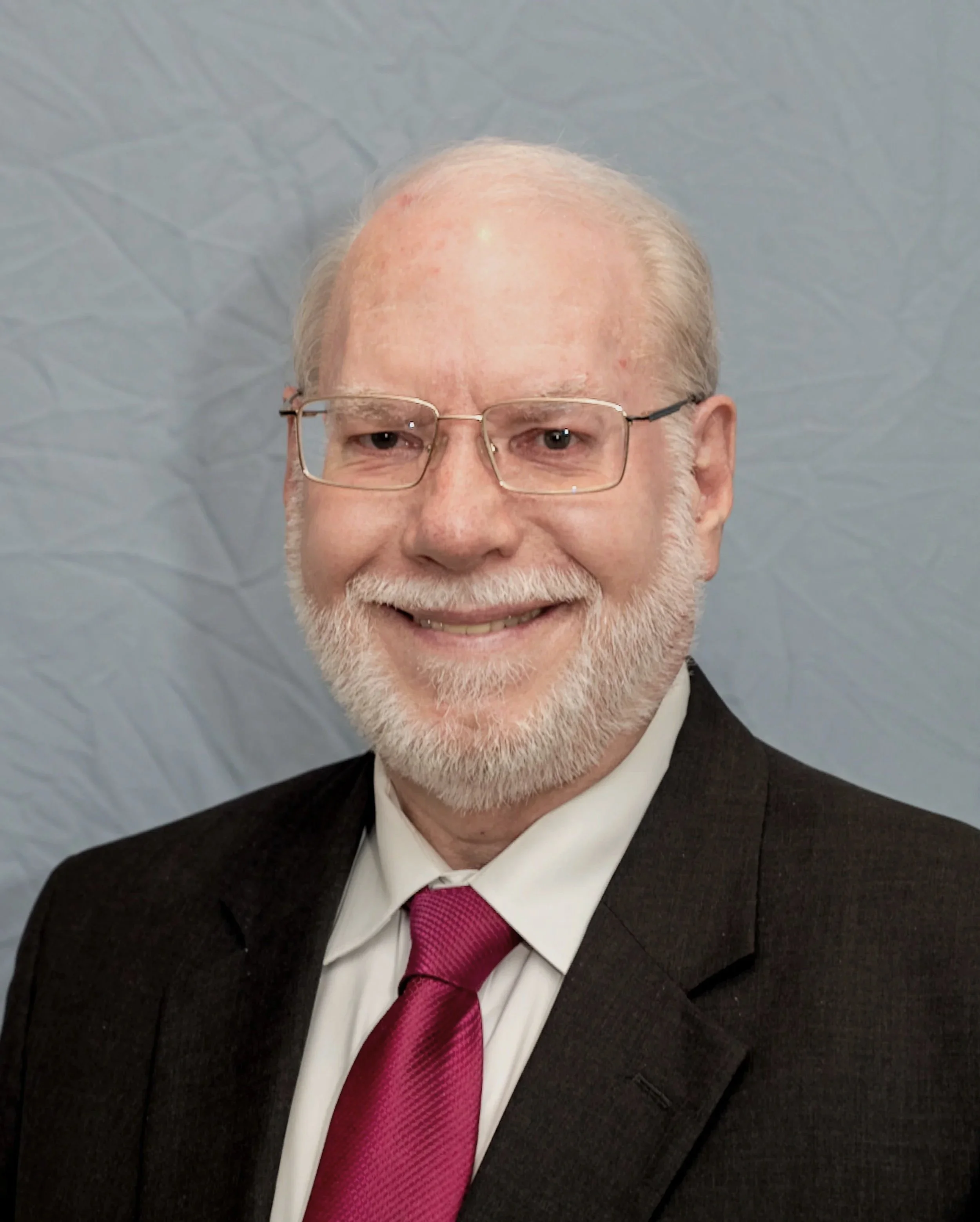 Close-up of an older man with glasses, white hair, and a beard wearing a black suit, white shirt, and magenta tie, smiling in front of a gray patterned background.