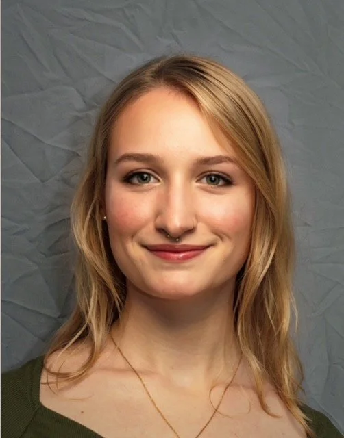 A young woman with blonde hair, blue eyes, and light skin smiling in front blue wall, wearing a green top, gold necklace, and facial piercings.