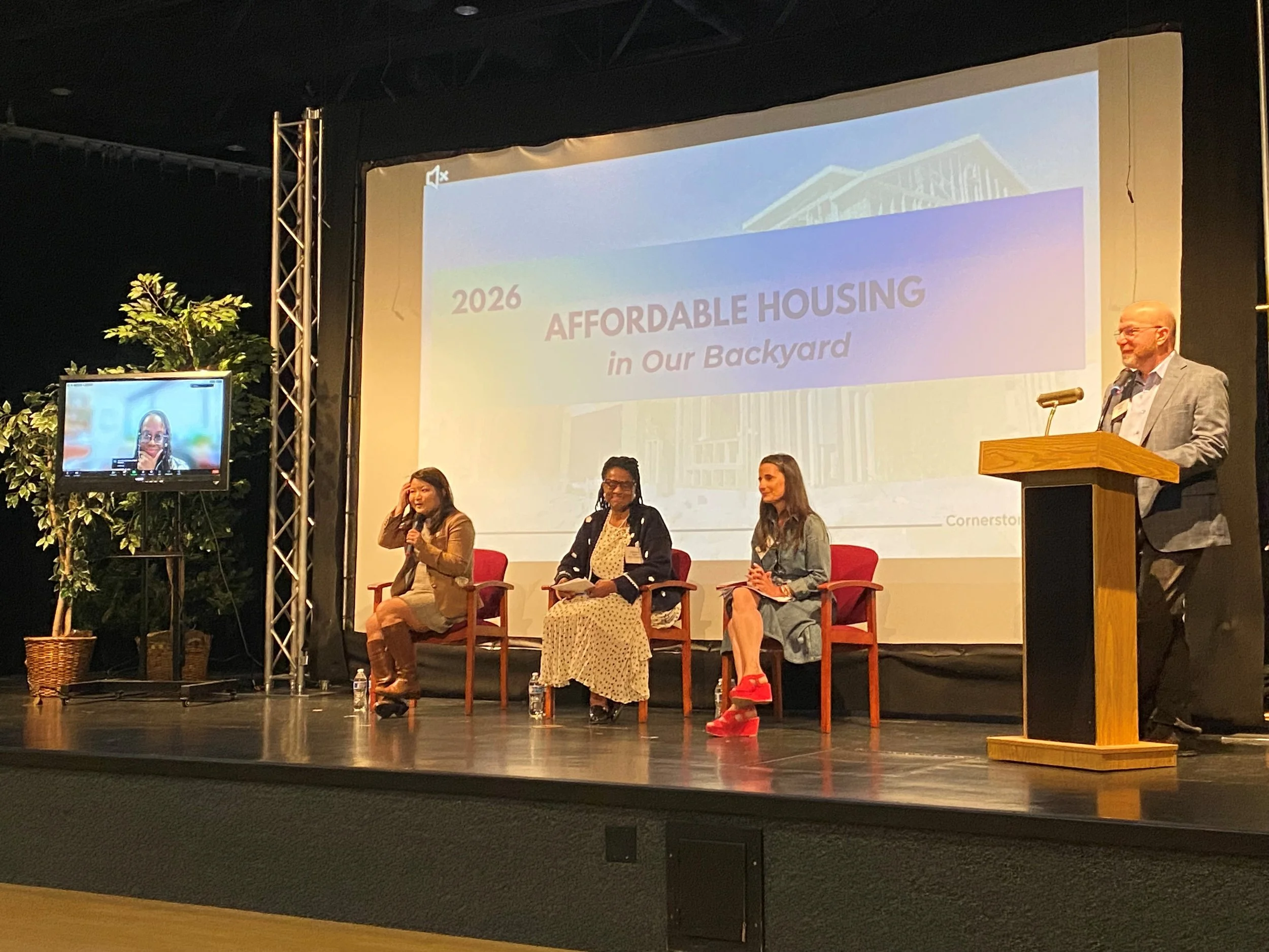 A panel discussion at a conference with four women and one man on stage. The man is standing at a podium, while the women are seated in chairs. A presentation slide behind them reads '2026 Affordable Housing in Our Backyard.' There is a large plant on the left side and a monitor displaying a woman with a headset in the foreground.