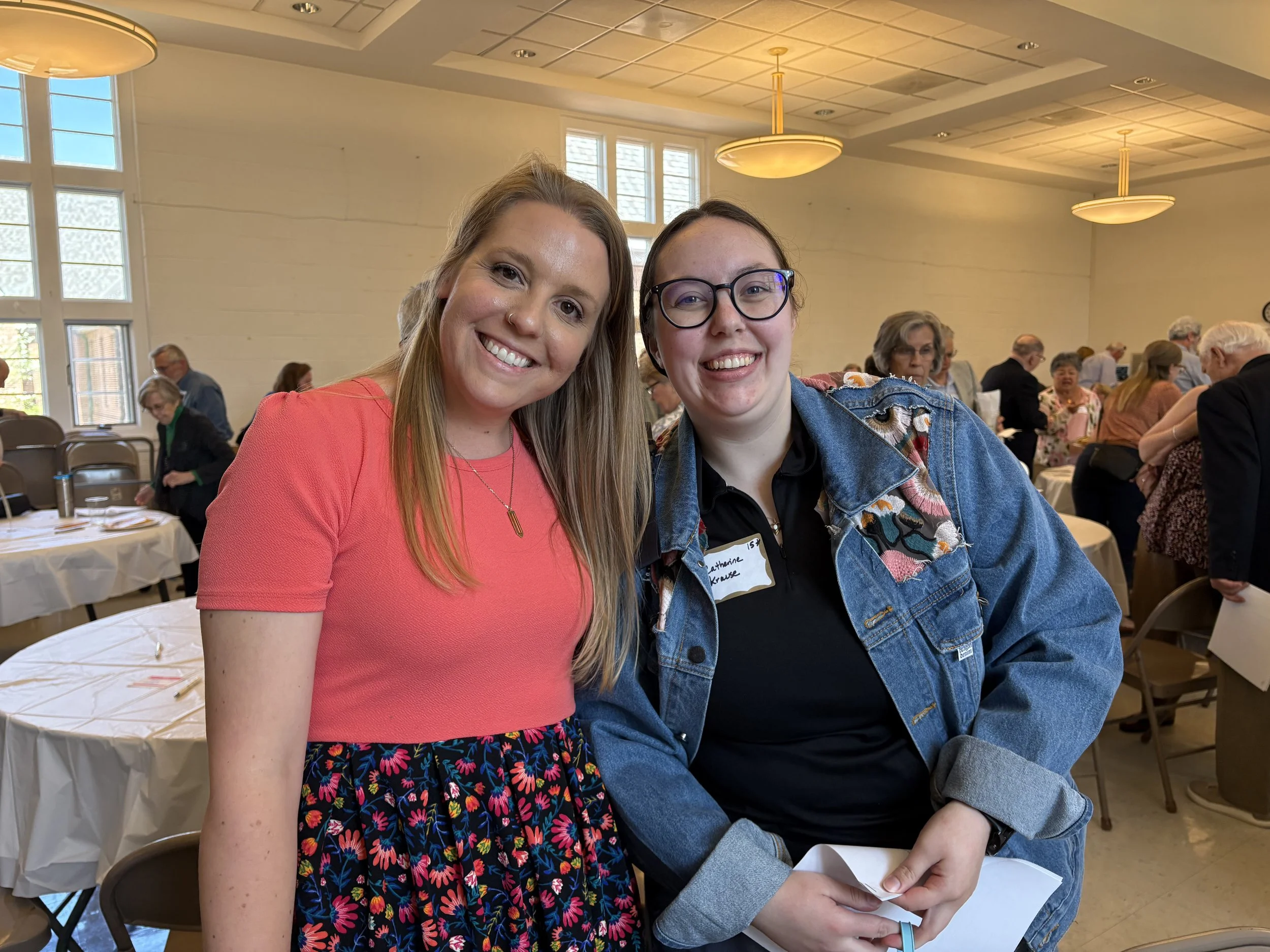 Two women smiling at the camera at a social event, sitting at a table with other people in the background. One woman wears a red shirt with a floral skirt, the other wears glasses and a denim jacket.