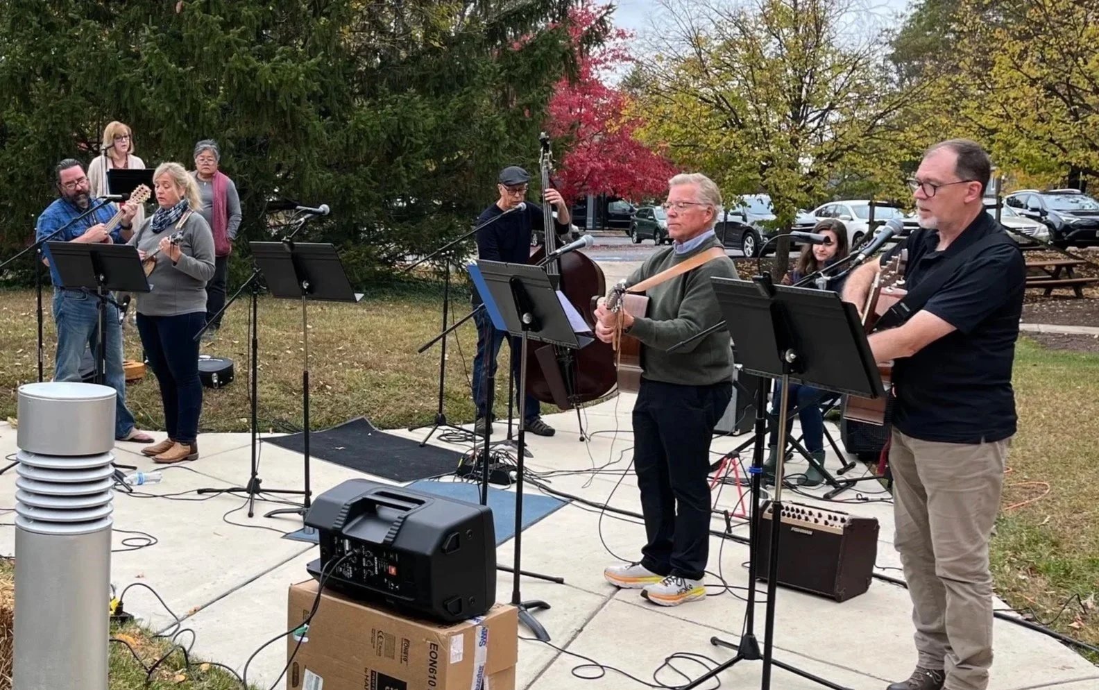 A group of musicians performing outdoors on a patio with trees and parked cars in the background. The group includes two vocalists, a guitarist, a bassist, and a drummer. Some members are reading sheet music, and microphones are set up for sound amplification.